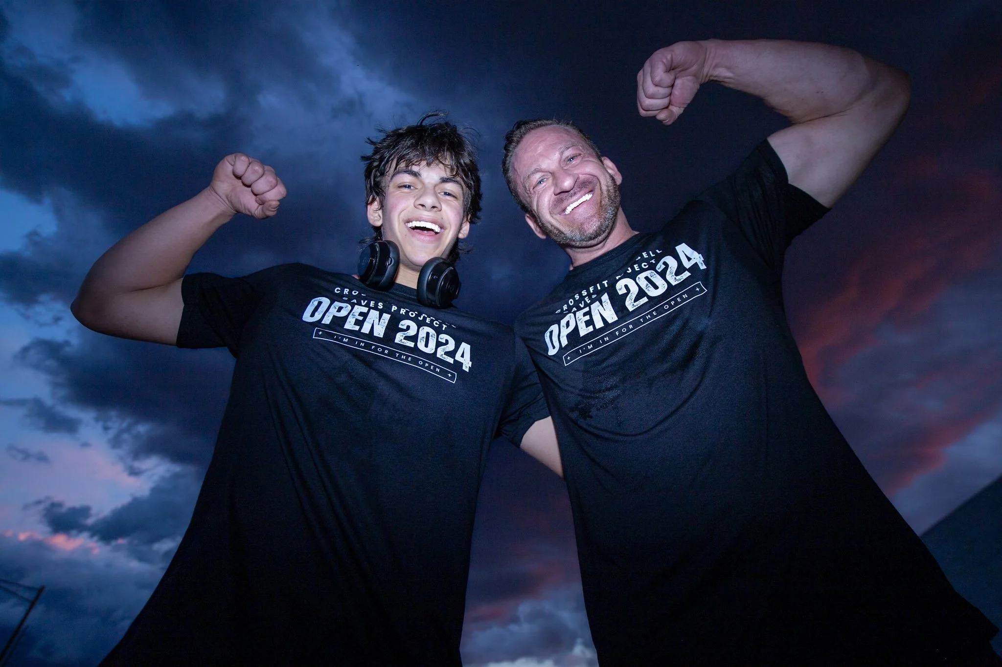 Two men smiling and flexing their biceps, wearing black T-shirts that say 'Open 2024' and 'CrossFit Project,' standing outdoors with a dark cloudy sky behind them.
