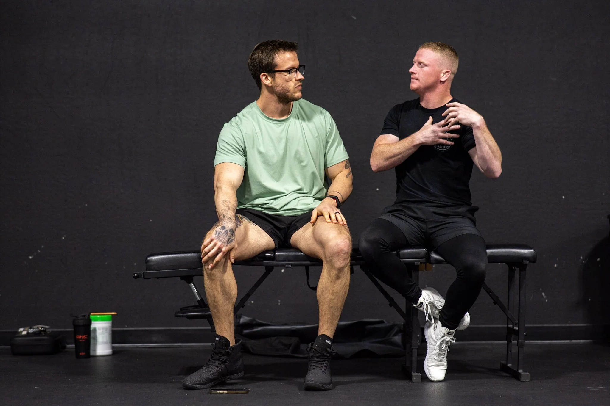 Two men sitting on therapy tables, engaged in conversation, against a black background. One is wearing a green shirt and shorts, with tattoos on his arm, and has his hand resting on his knee. The other is wearing a black shirt and leggings, with his hand on his chest, appearing to speak.