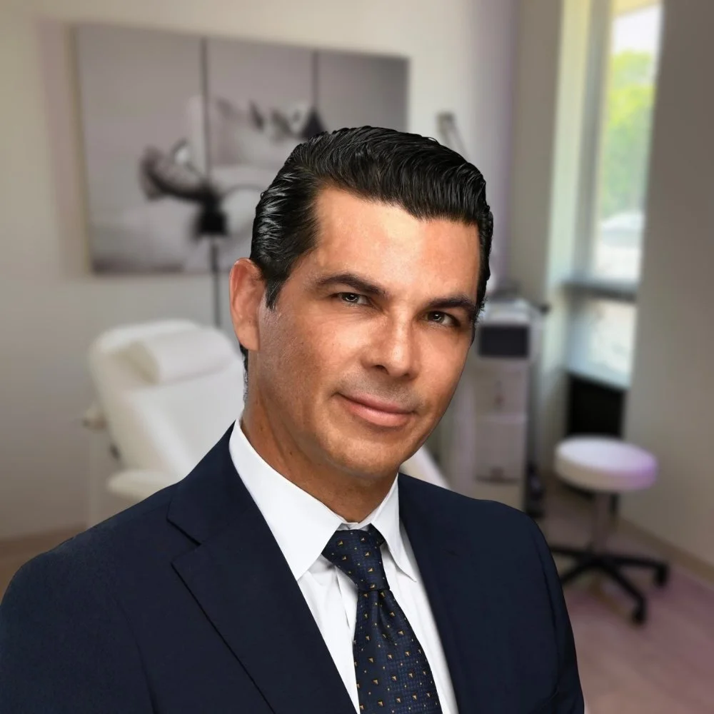 A man in a navy suit and tie in a medical office with an examination chair and medical equipment in the background.