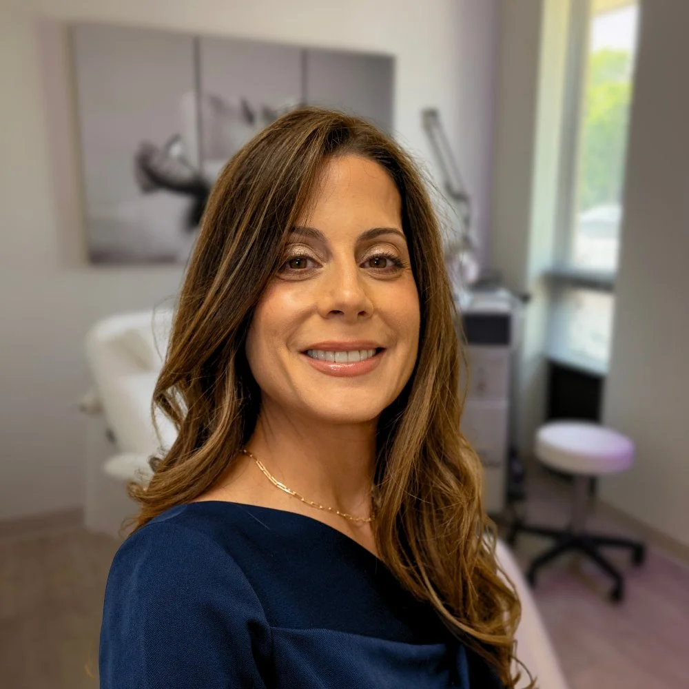 A woman with long brown hair smiling in a medical or clinical setting, with a dentist's chair and medical equipment in the background.