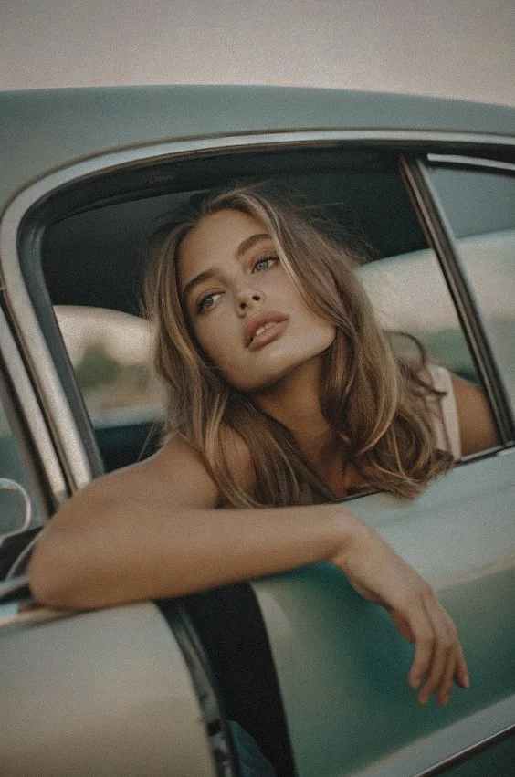 Picture of a young woman in a car with the window rolled down and her arm hanging over the edge looking wistfully to the distance