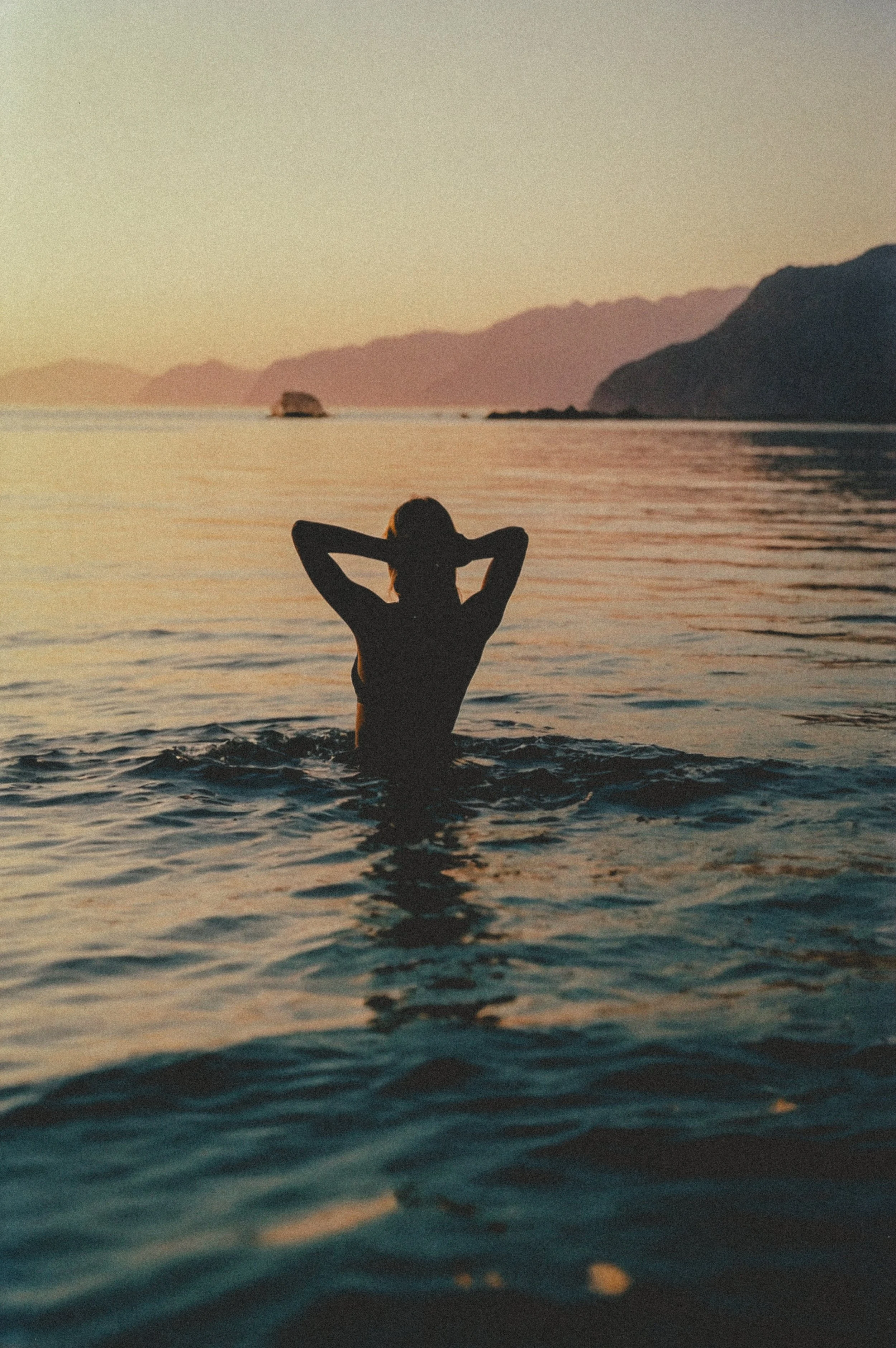 Woman is wading out into the ocean with a beautiful sunset and rock cliffs and mountains off into the distance