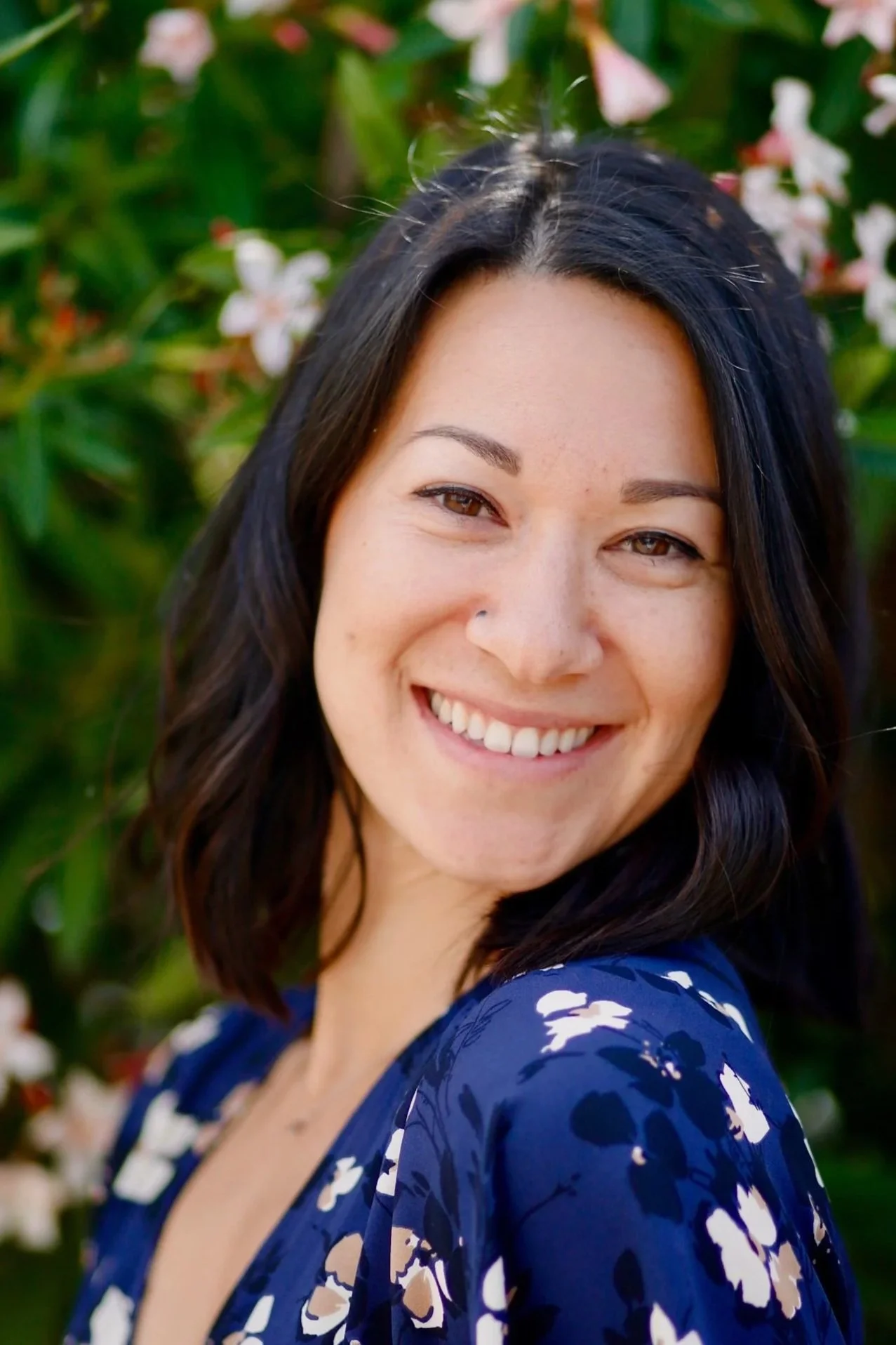 A woman with black hair smiling outdoors, wearing a blue floral dress, with green plants and pink flowers in the background.