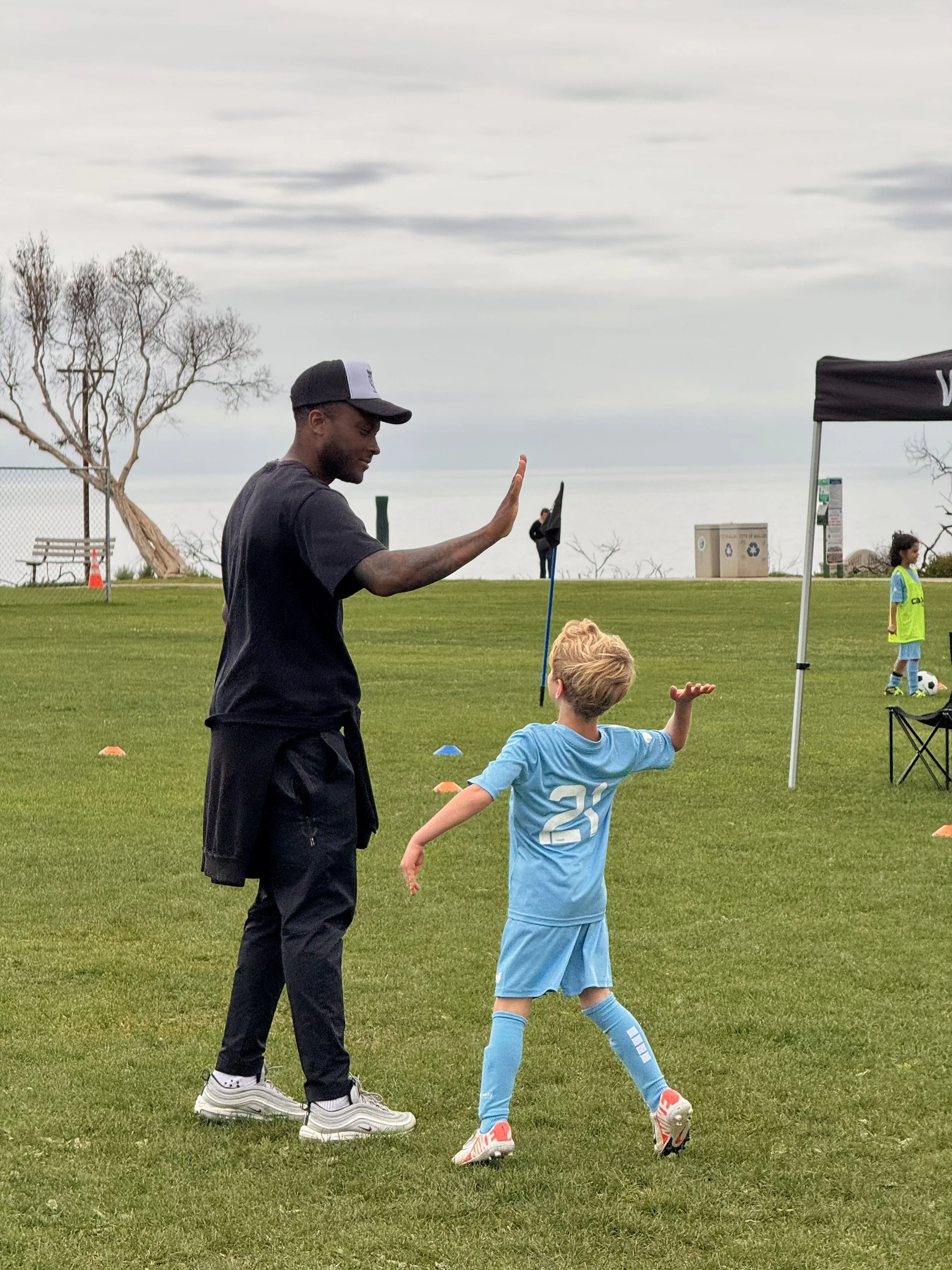 A soccer coach high-fiving a young player in a light blue jersey on a grassy field under a cloudy sky.