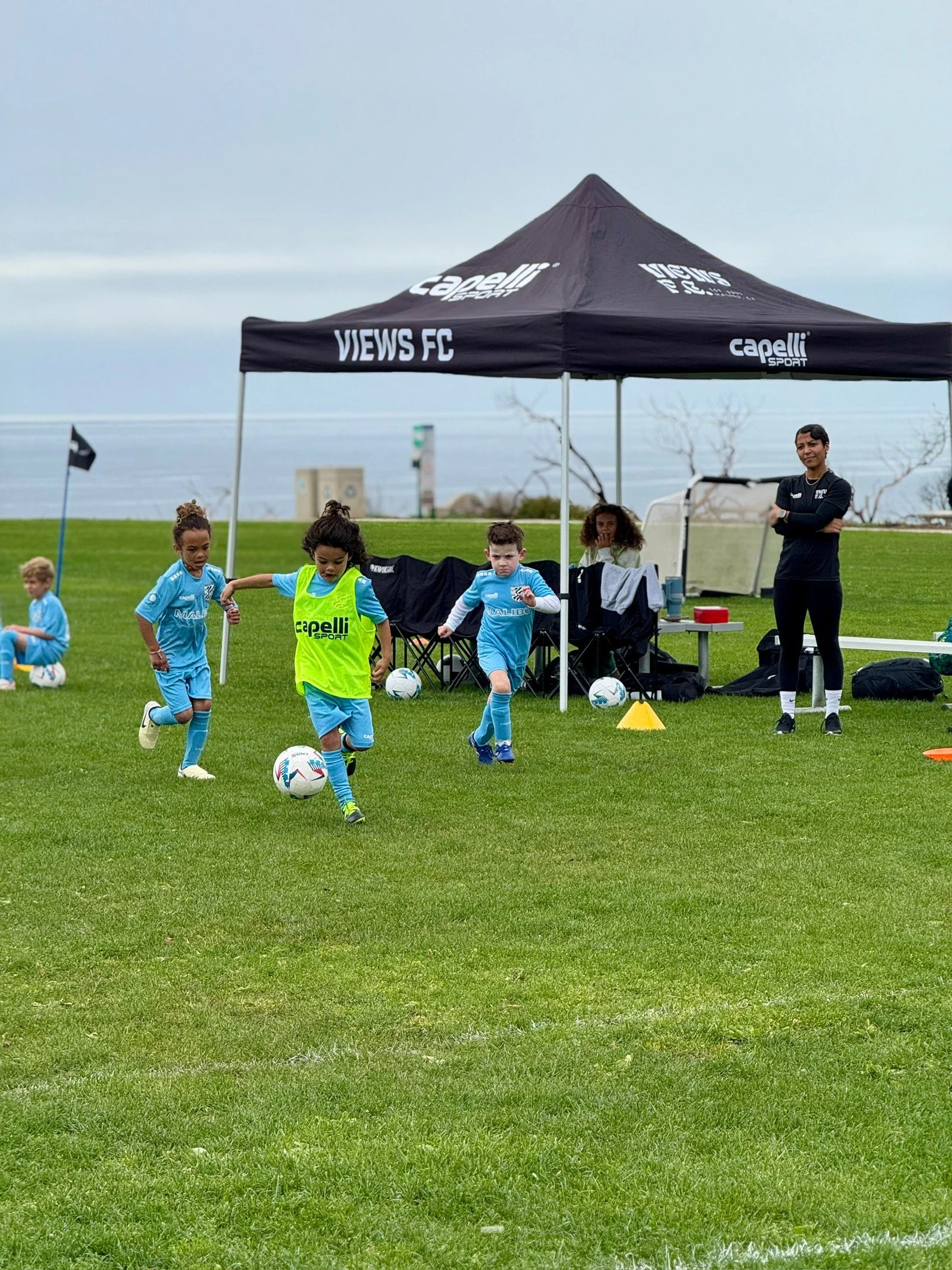 Children playing soccer on a grass field under a "Capelli Sport" tent with "VIEWS FC" written on it. A coach and spectators are watching on the sidelines. The ocean is visible in the background.