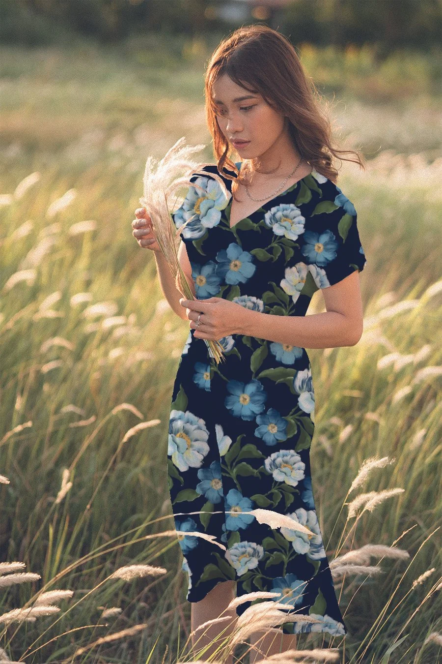 A young woman with light brown hair wearing a navy dress with a floral pattern holding a small bouquet of dried flowers in a grassy field during sunset.