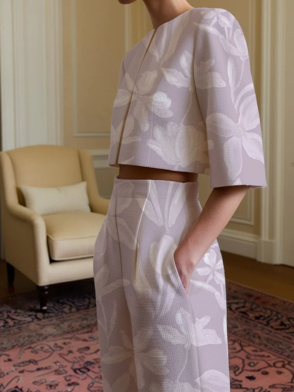 A woman wearing a light-colored, floral-patterned, cropped jacket and matching high-waisted wide-leg pants, standing with her left hand in her pocket in a room with a cream armchair and beige walls.