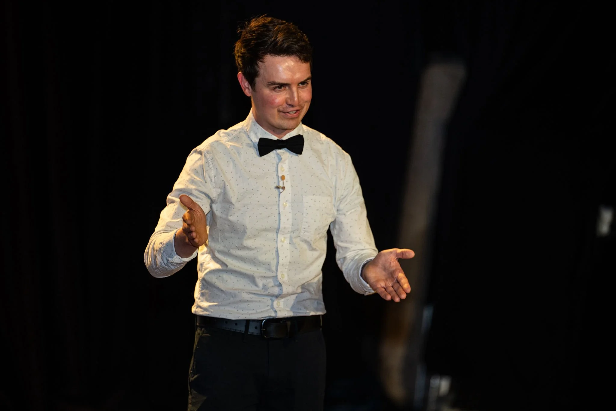 Ben Rush, wearing a white shirt with a black bow tie, gesturing with his hands, emceeing a Wisconsin Science Festival event