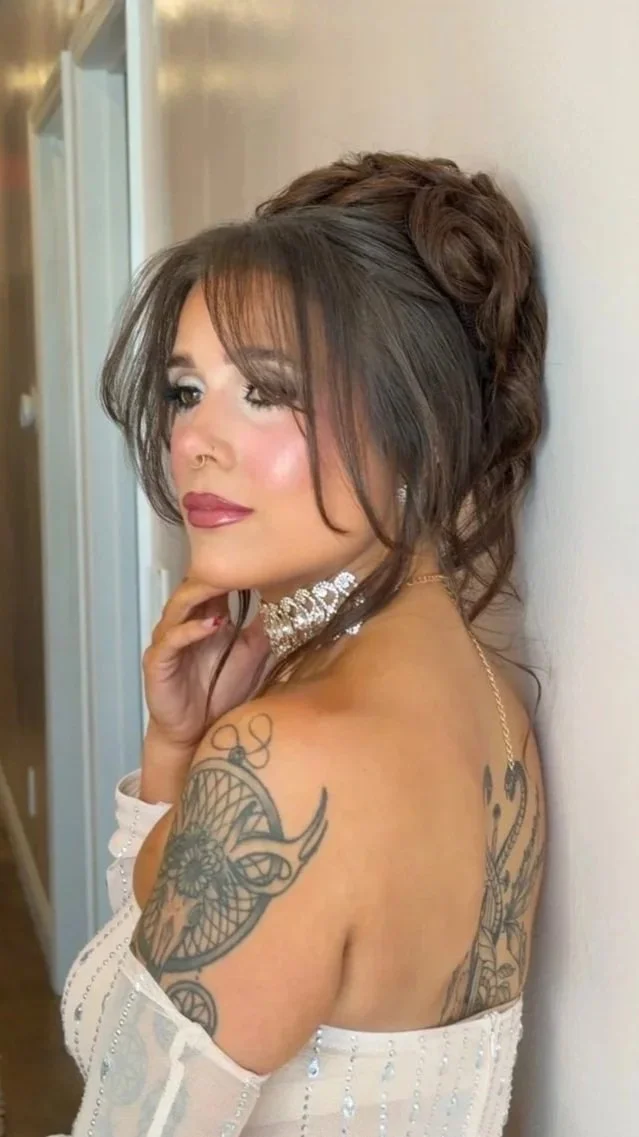 A woman with brown hair styled in a voluminous updo and soft bangs, wearing a white off-shoulder dress with sequins, adorned with jewelry including a large choker and earrings, standing against a light-colored wall. She has tattoos on her shoulder an