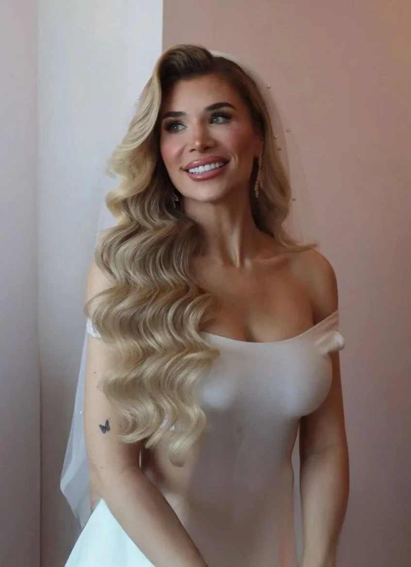 A woman with long wavy blonde hair, wearing a white off-shoulder dress, smiling and looking to her right. She has earrings and a small tattoo of a butterfly on her left arm.