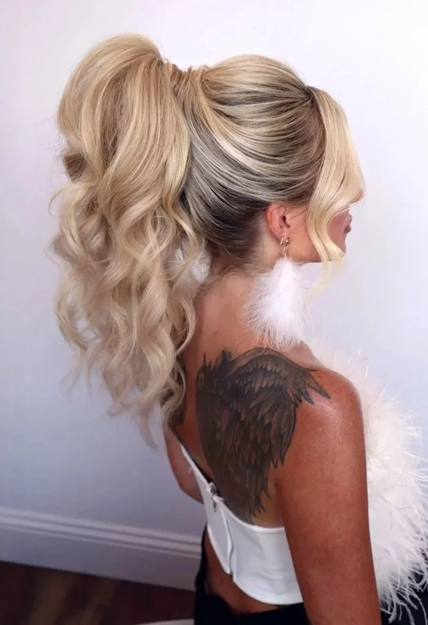 Side view of a woman with long, voluminous, blonde curly hair styled in a high ponytail, wearing white feather earrings and a white top, with a black feather tattoo on her shoulder.