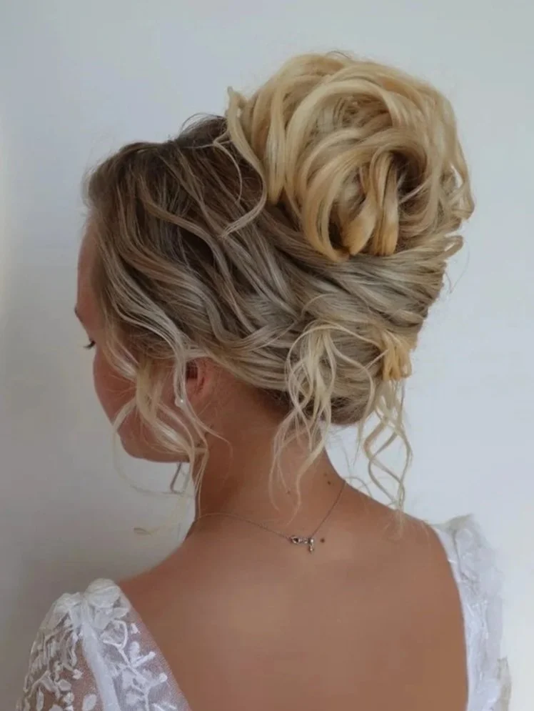 Side view of a woman with blonde, curly hair styled in an updo, wearing a white dress with lace details and a delicate necklace.
