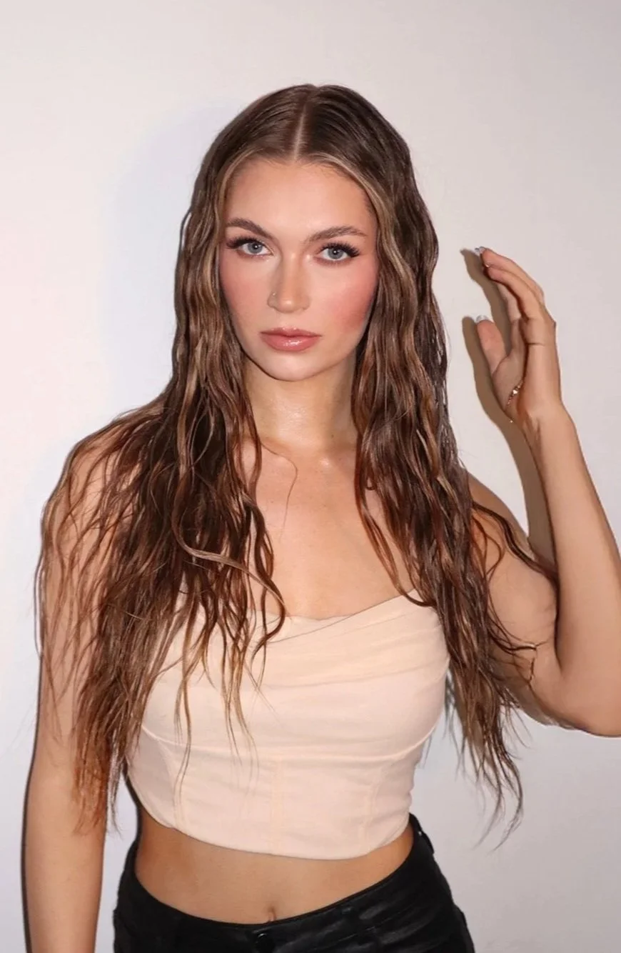 A young woman with long wavy brown hair, wearing a strapless beige top and black pants, posing against a plain white background.