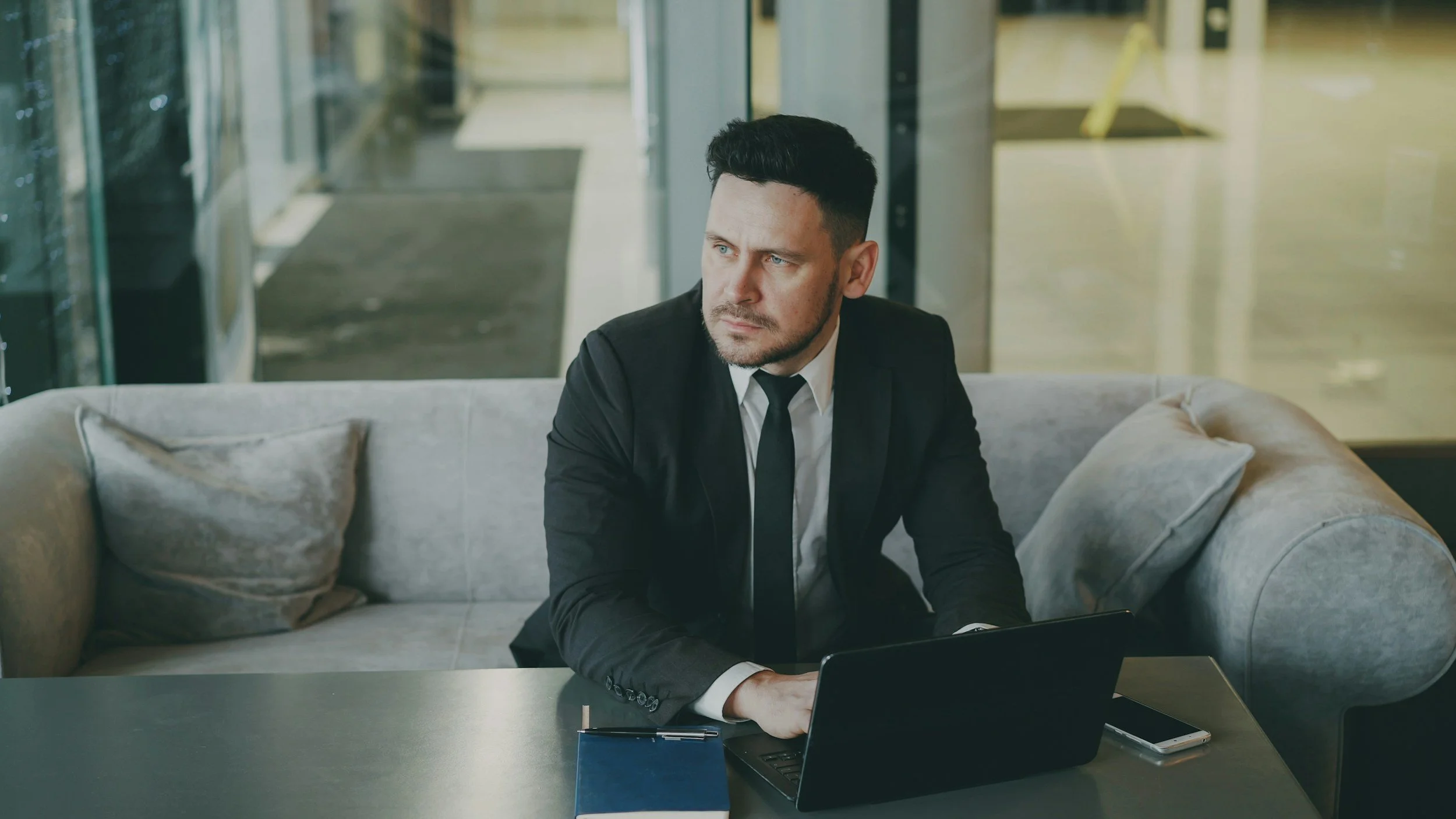 A man in a black suit with a white shirt and black tie sits on a sofa in a modern office lobby. He has short dark hair and a beard, and looks to the side with a serious expression. On the table in front of him are a blue notebook, a laptop, and a smartphone.