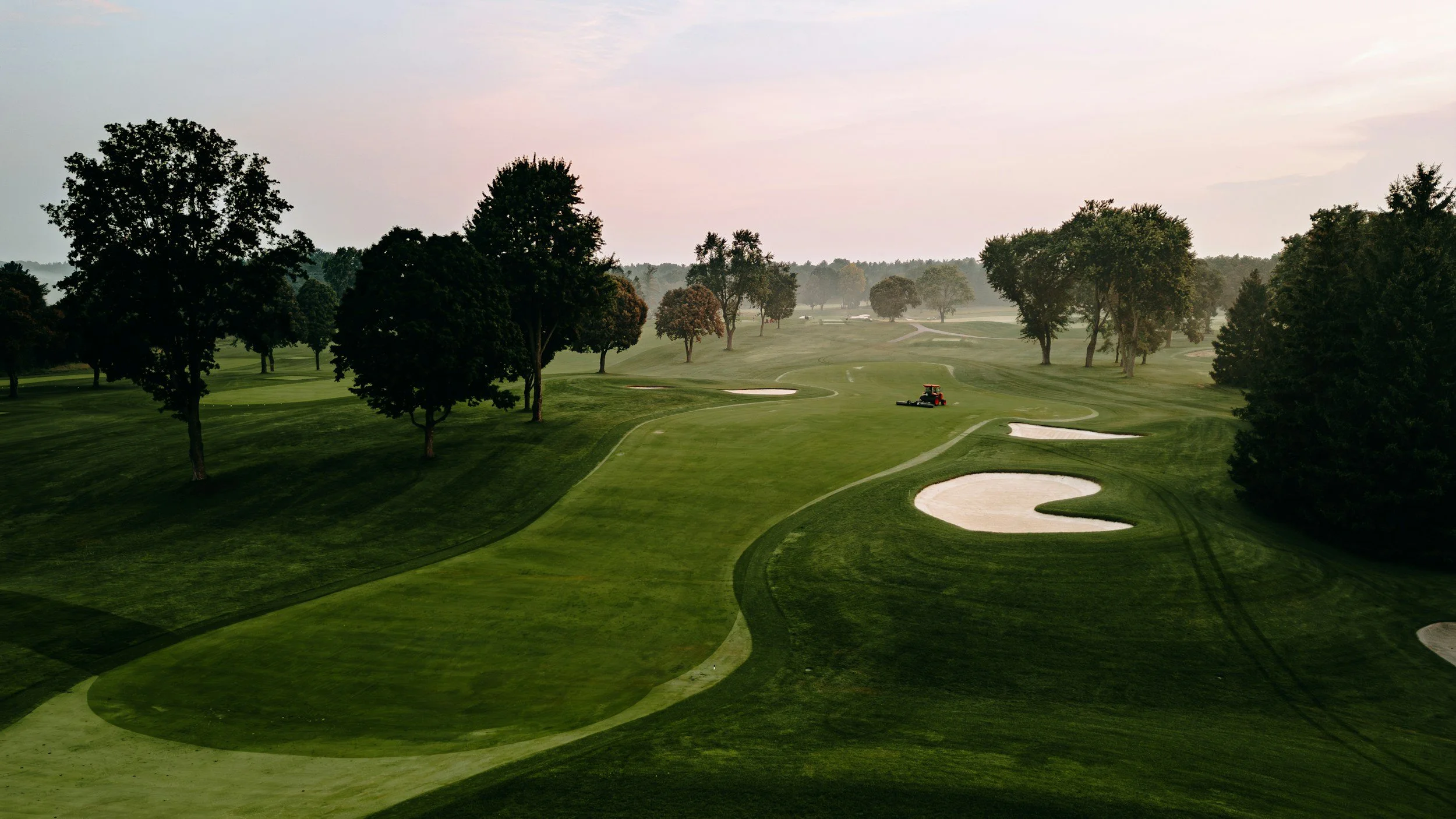 A golf course at dawn with green fairways, sand bunkers, and trees lining the course. A golf cart is visible in the distance.