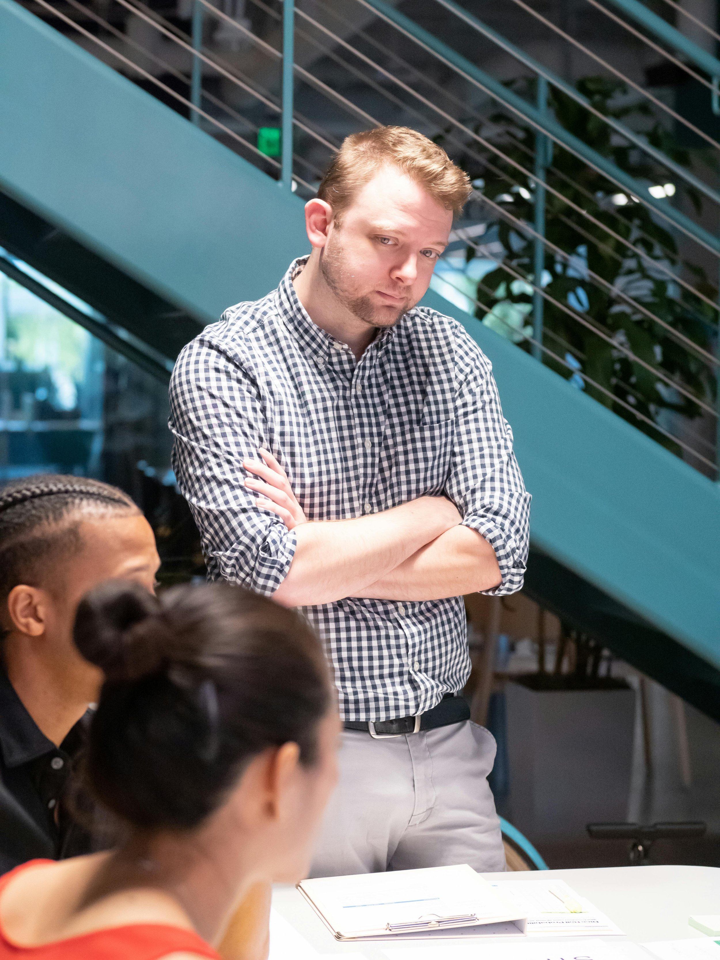 Man with crossed arms looking at seated women in a meeting or discussion in a modern office space.