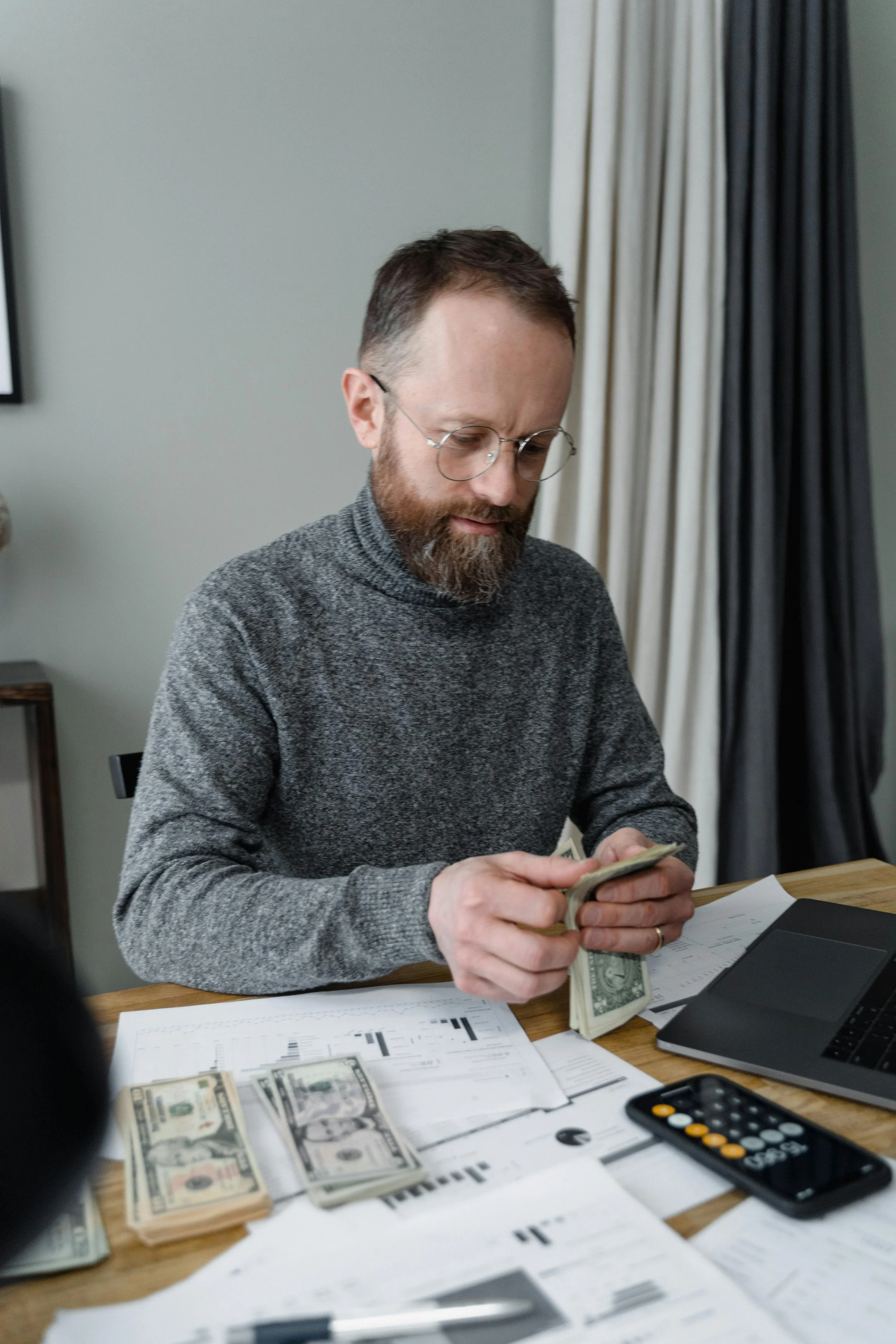 Man with glasses counting cash at a desk with documents, a calculator, and a laptop.