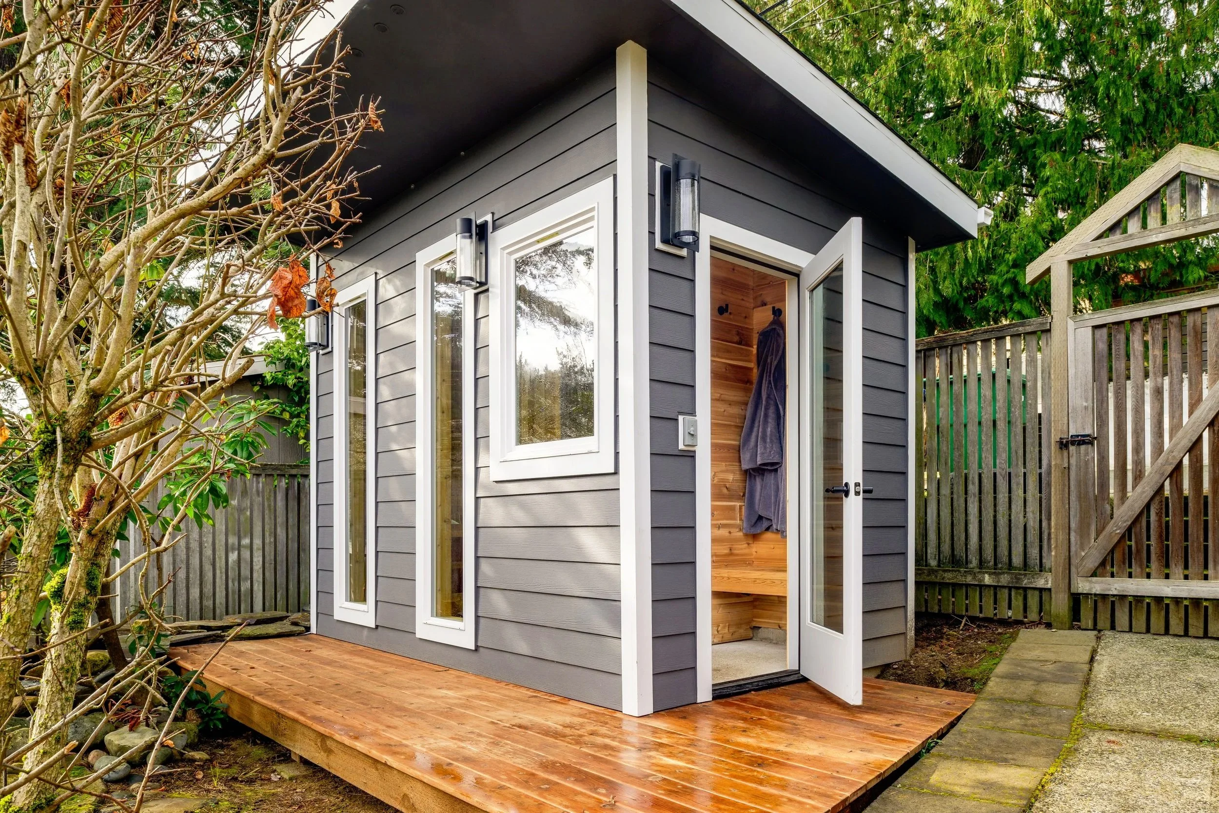 A small modern gray outdoor shed with wooden flooring on a patio, surrounded by a wooden fence, with a tree in the foreground and greenery in the background.
