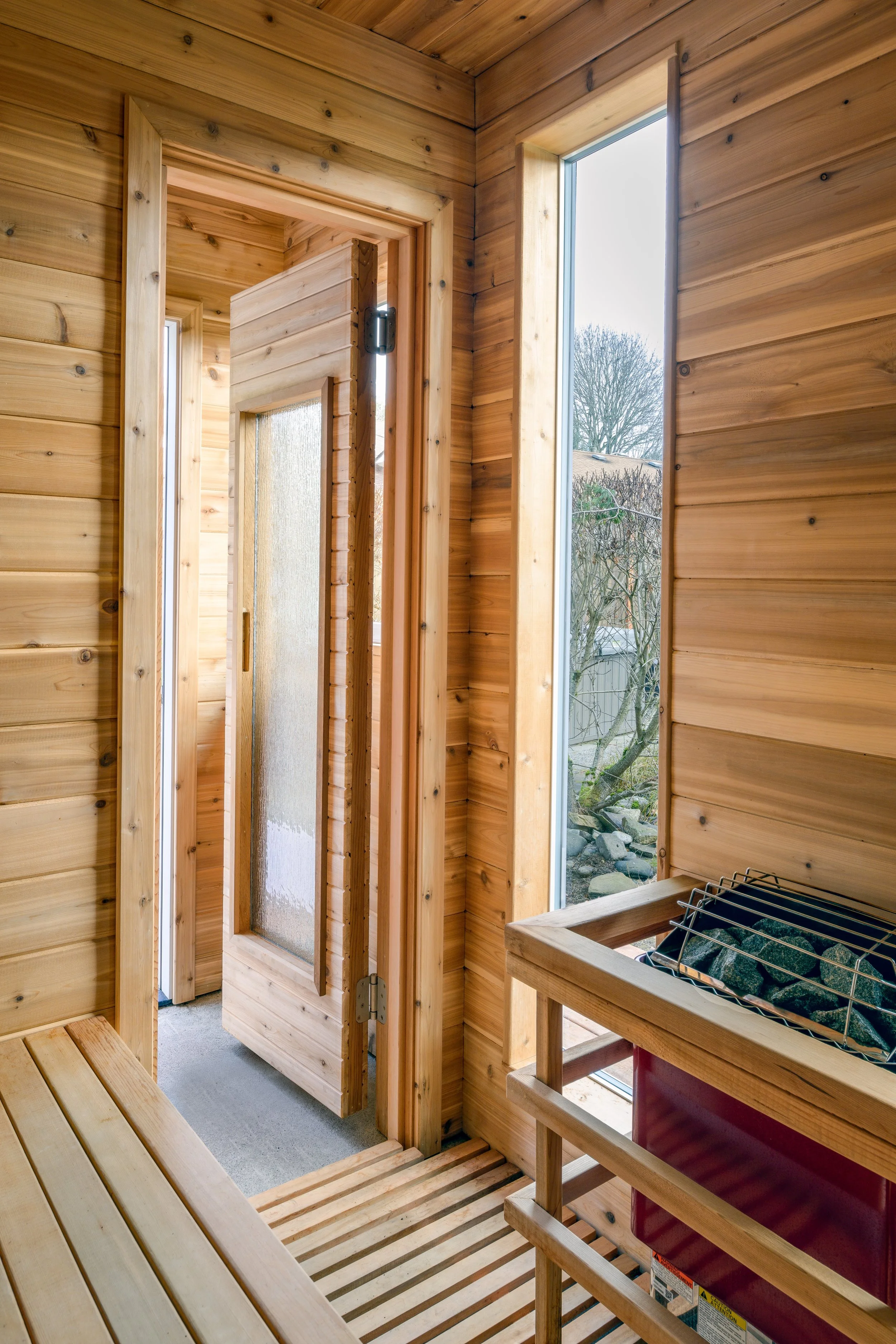 Interior view of a wooden sauna with a door, large window, and a heater with stones.