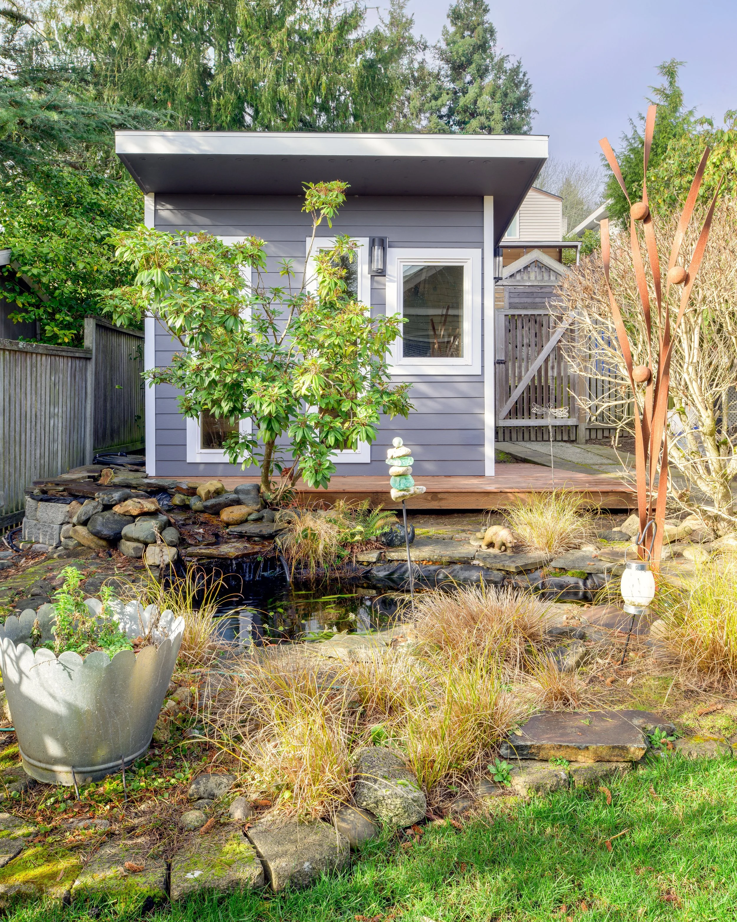 A small house with gray siding and white window trim. A wooden deck in front of the house with garden plants, a tree, a rock waterfall feature, and decorative items like a stack of stones and a lantern. A fence and trees are in the background.