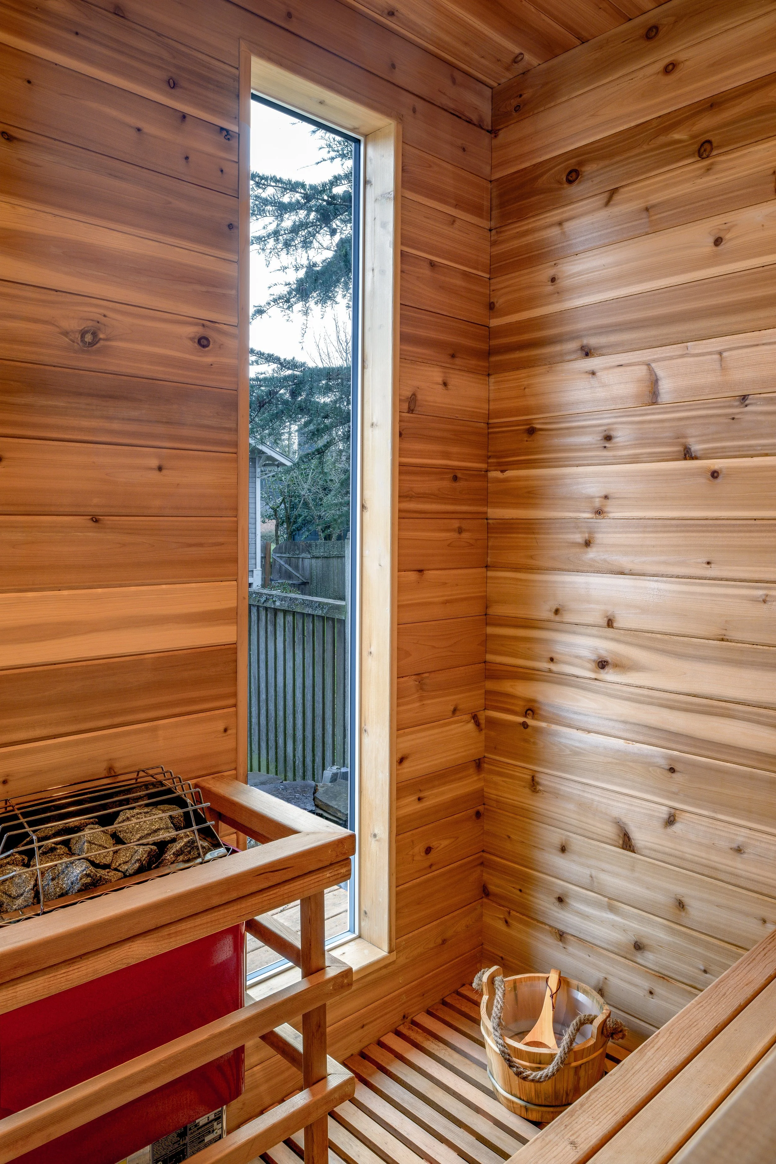 Interior of a sauna with wooden walls, a small window showing an outdoor fence and trees, a sauna heater with rocks, and a wooden bucket with a ladle.