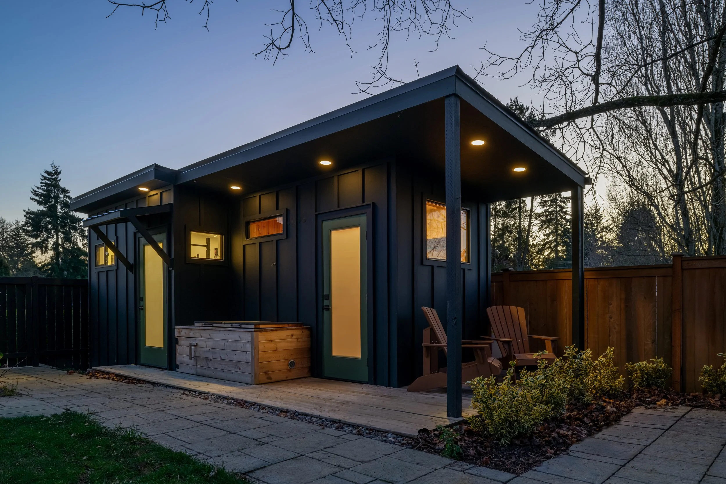 A modern small backyard building with a black exterior, yellow door and windows, illuminated by ceiling lights, surrounded by wooden fencing, with outdoor seating and plants, during sunset.
