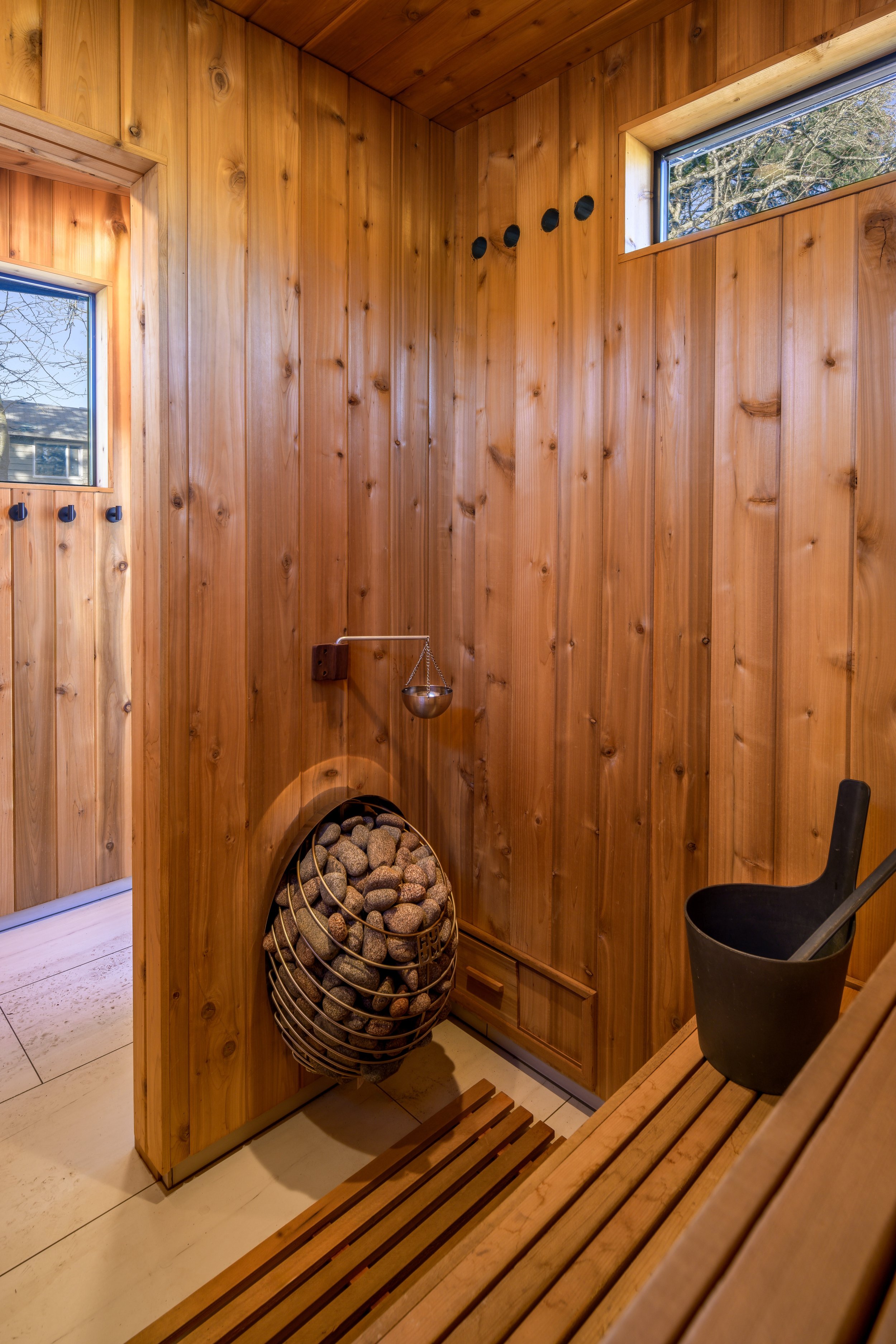 Wood-paneled sauna room with small windows, a bucket and ladle, rocks in a basket, and bench seating.