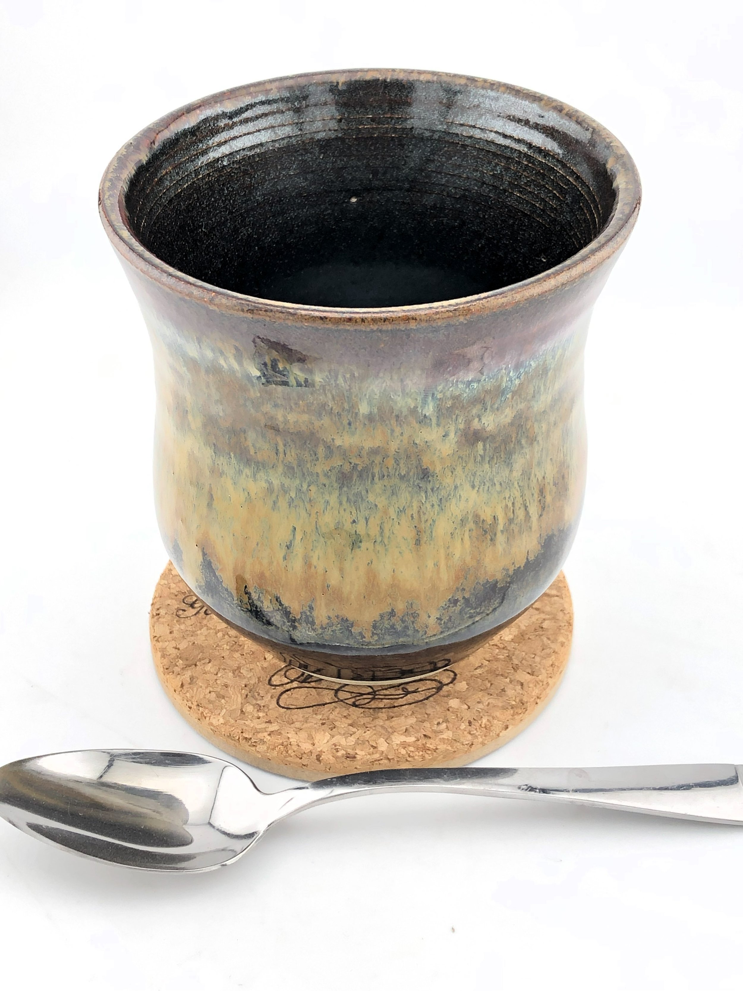 Ceramic cup with rustic glaze on cork coaster, next to a metal spoon.