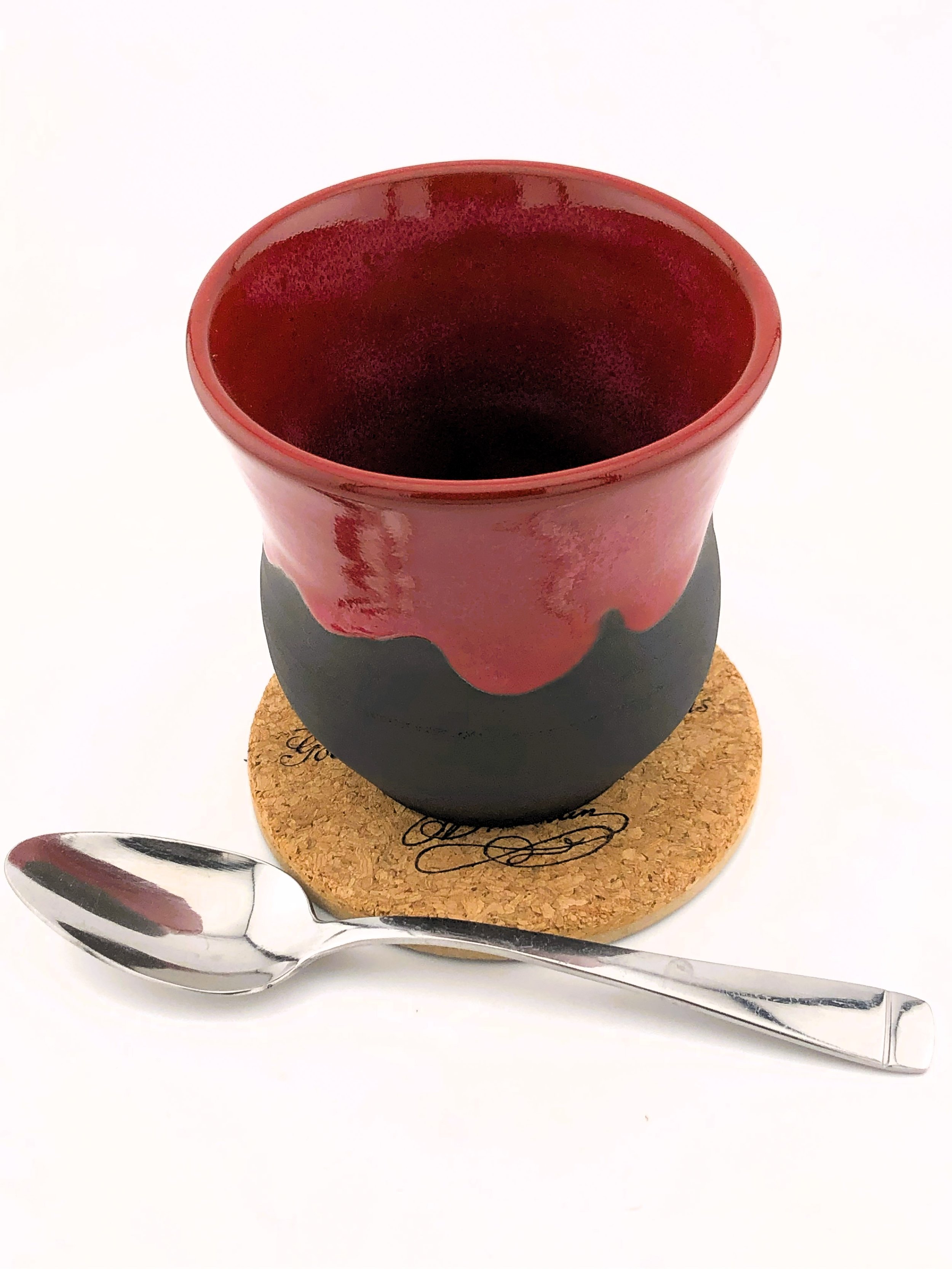 Red and black ceramic cup on cork coaster with silver spoon.