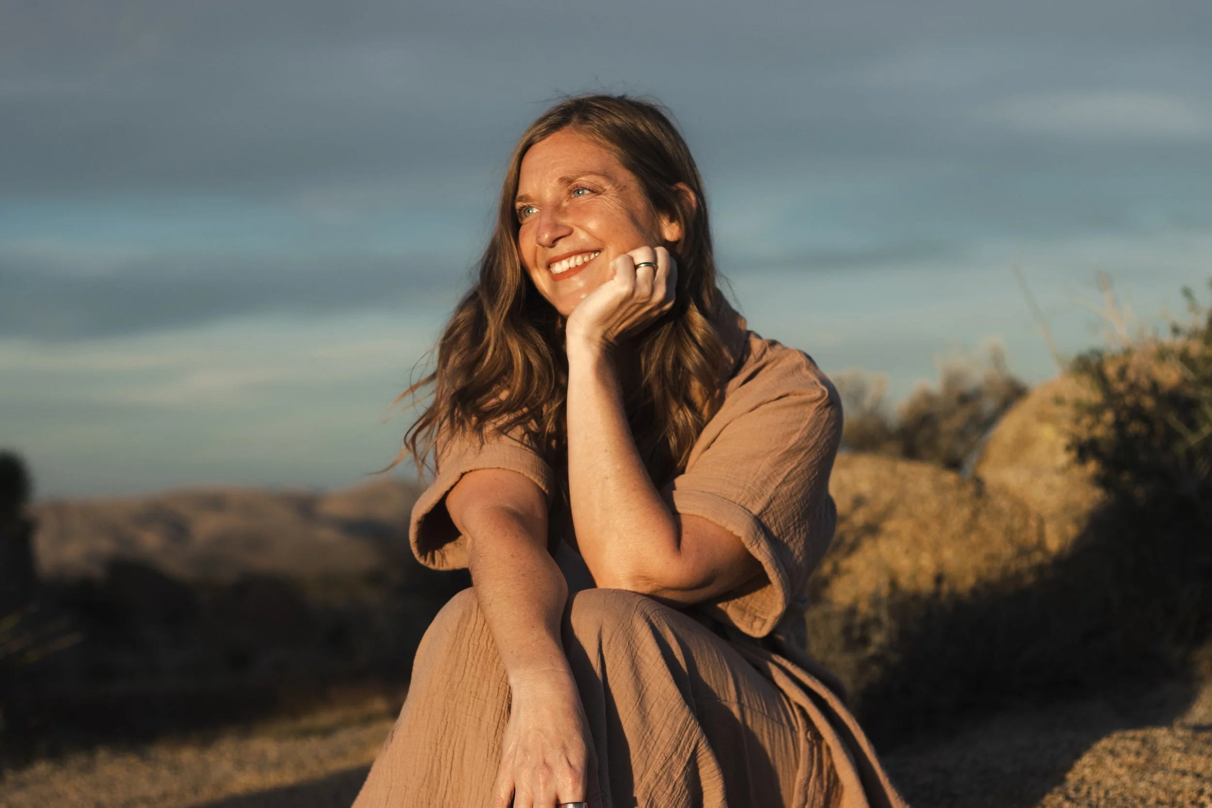 A woman with long brown hair smiling outdoors during sunset, sitting on a rocky landscape in Joshua Tree with a cloudy sky in the background.