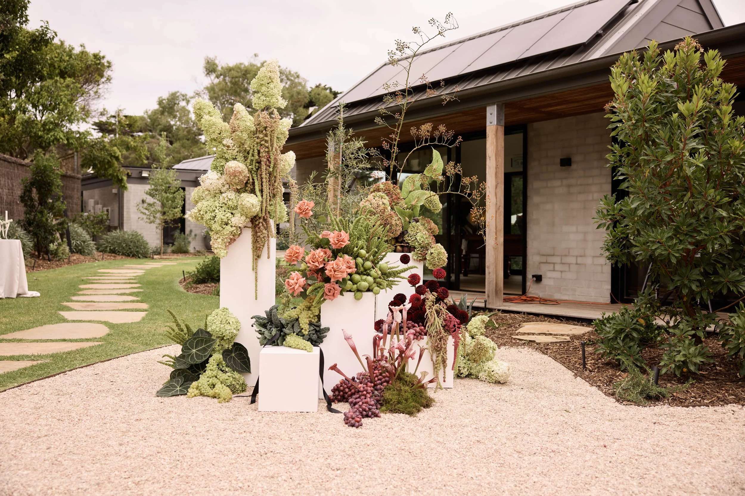 Outdoor space with garden and floral arrangements on white pedestals in front of a house with a covered porch, brick wall, and glass door.