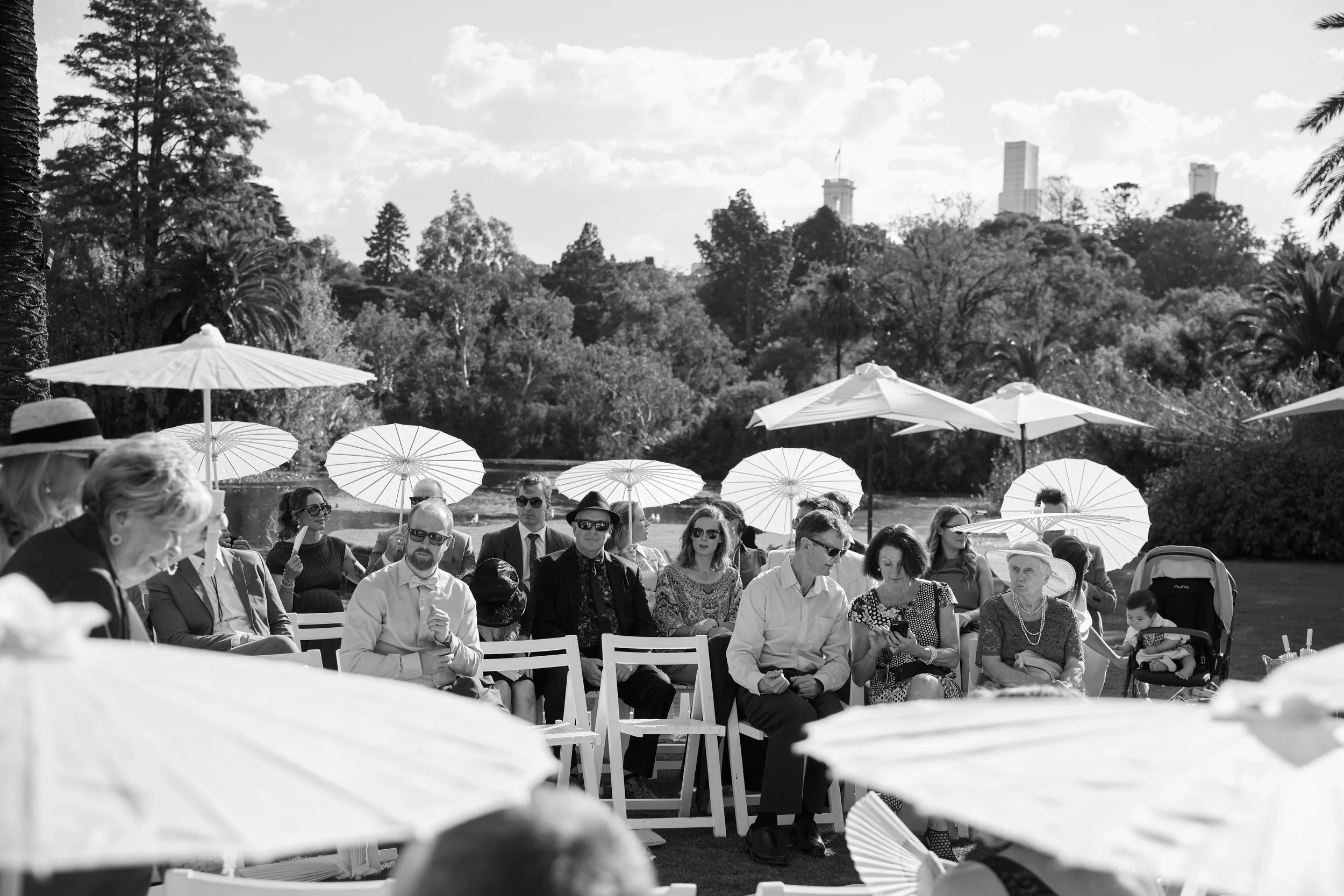 Black and white photo of a group of people sitting outdoors on chairs, with umbrellas overhead, near a body of water and trees, possibly at a formal event or gathering.