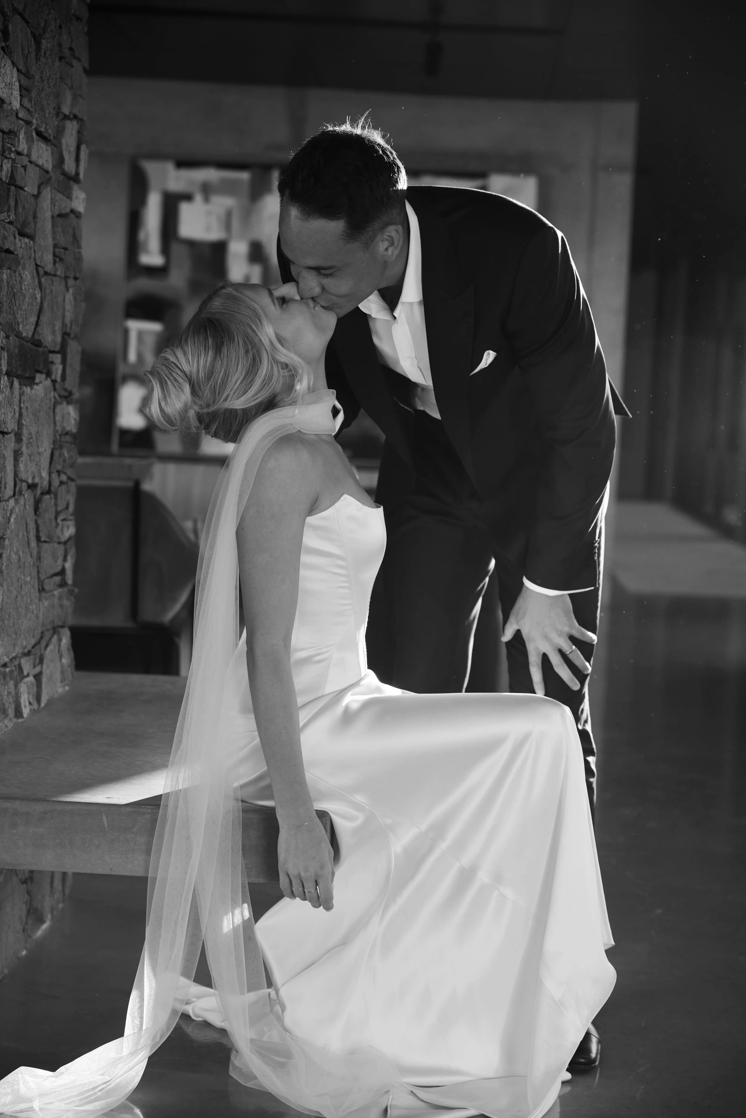 A black and white photo of a bride and groom sharing a kiss, with the groom leaning over the bride who is sitting on a bench. The bride is in a strapless wedding dress with a veil, and the groom is dressed in a dark suit and white shirt, indoors with a rustic stone wall on the left side.