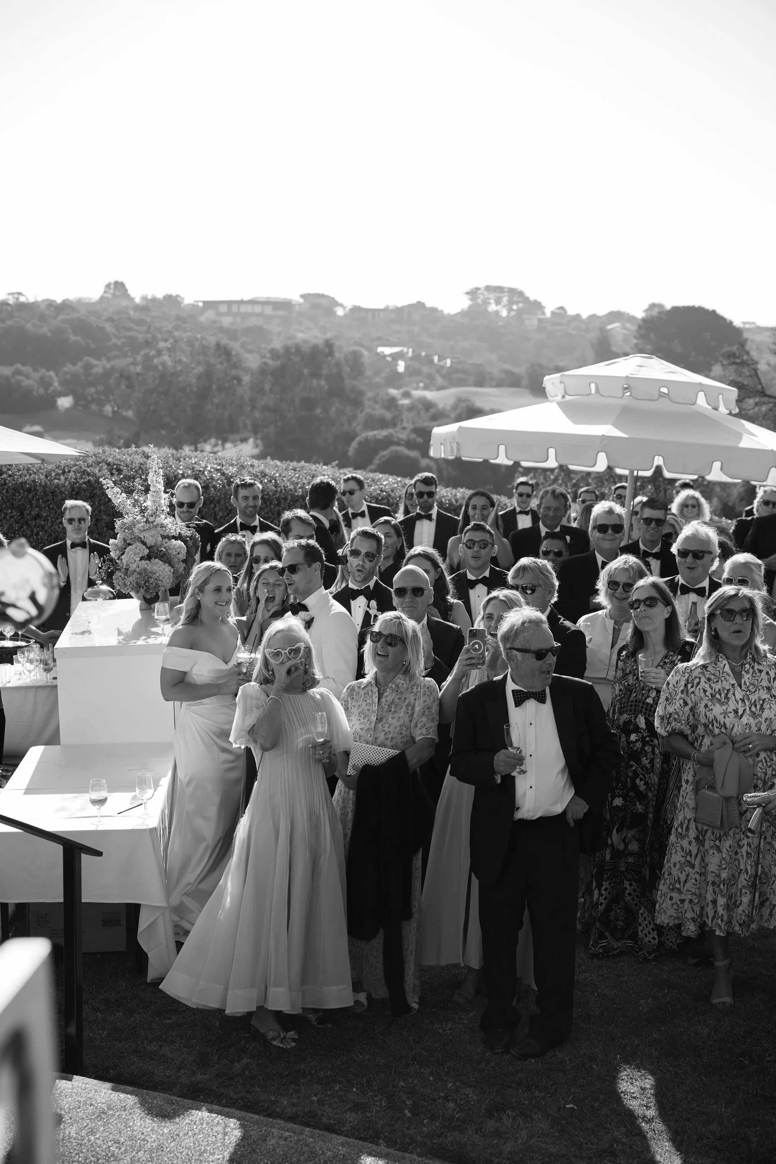 Guests dressed in formal attire, including tuxedos and elegant dresses, gathered outdoors for a celebration or event under umbrellas, with scenic landscape in the background.
