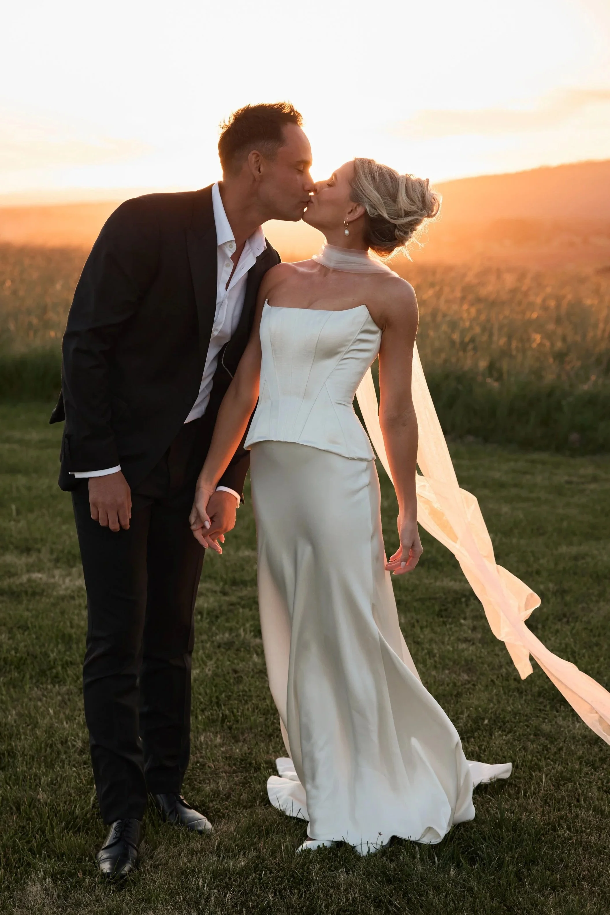 A newlywed couple sharing a kiss at sunset in a grassy field, with the groom in a black suit and the bride in a white wedding gown.
