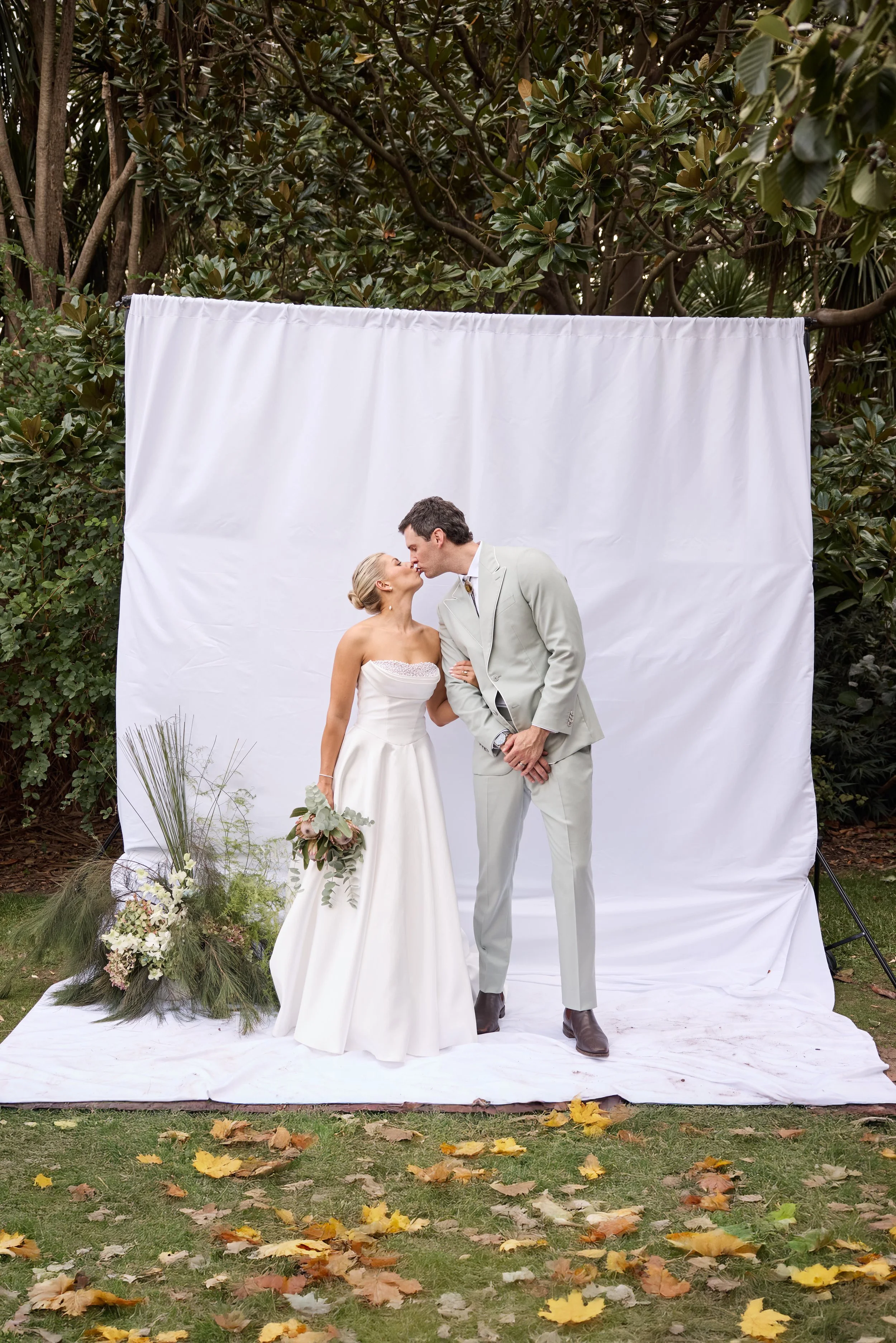 A couple in wedding attire kissing outdoors, standing in front of a white backdrop with greenery around, with fallen autumn leaves on the grass.