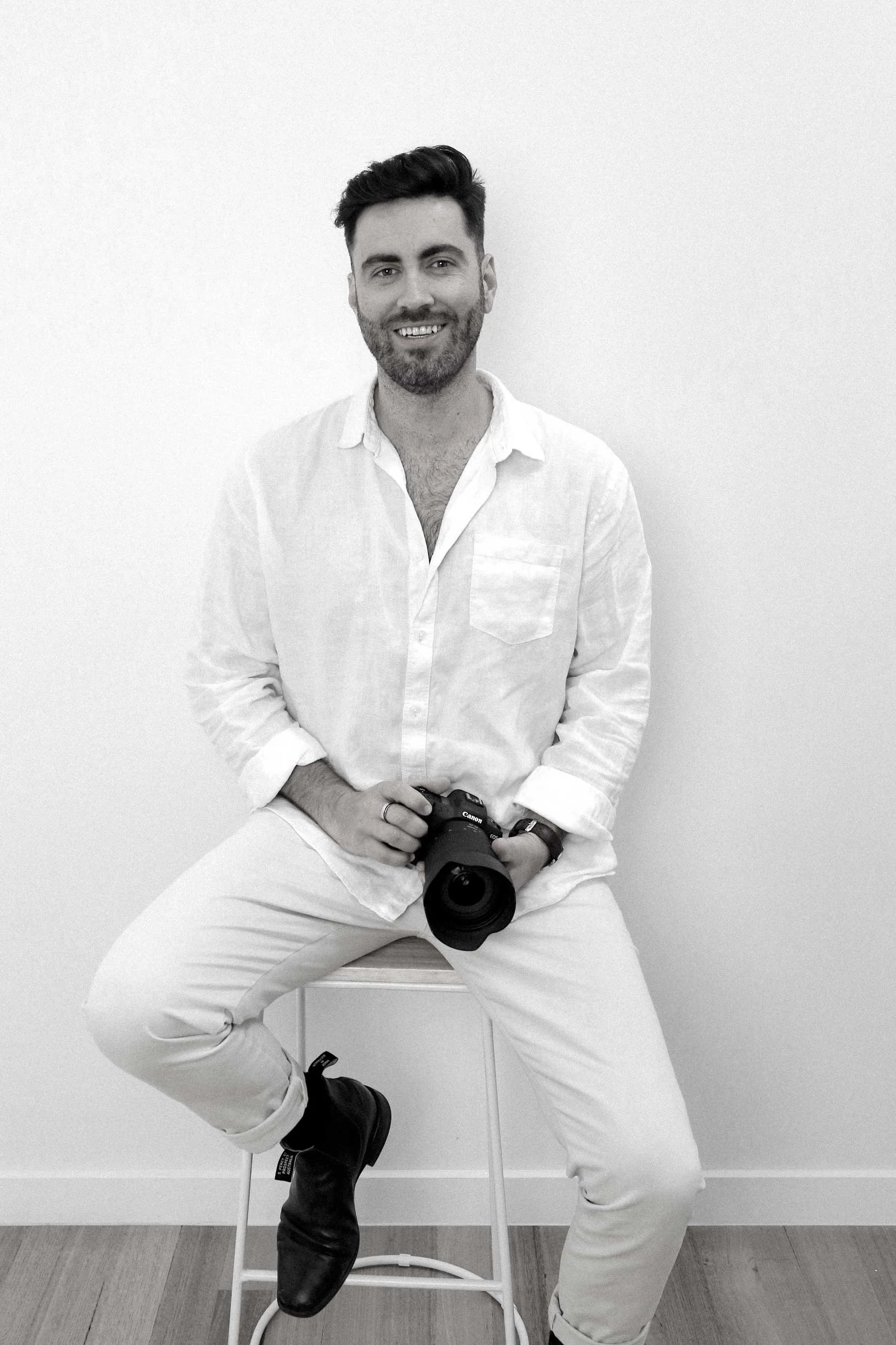 A man with dark hair and a beard, wearing a white shirt and white pants, sitting on a stool holding a camera, smiling, against a plain white wall.