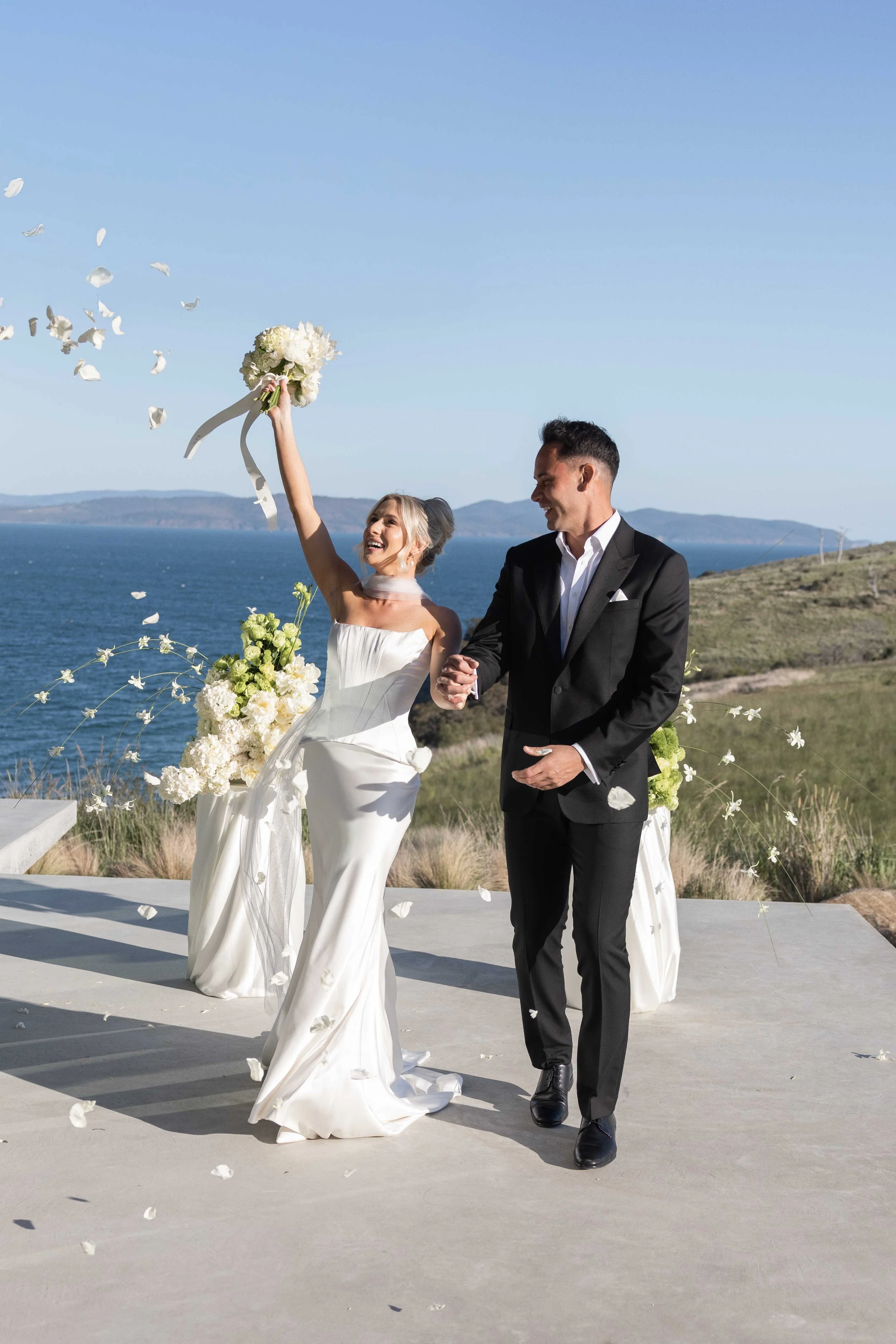 A wedding couple celebrating outdoors near the ocean, with the bride in a white dress holding a bouquet and the groom in a black suit. White flower petals are falling.