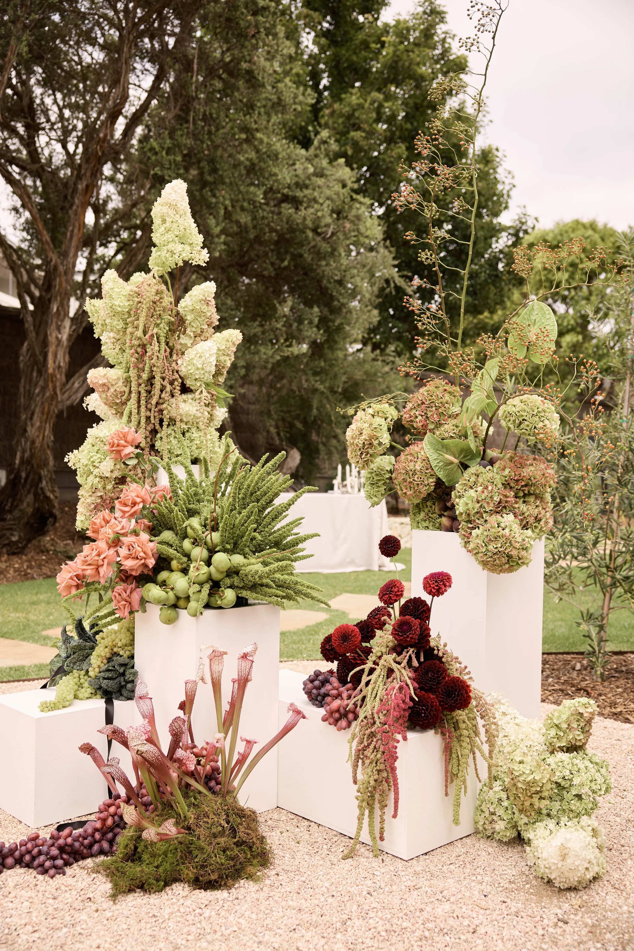 Elegant outdoor floral arrangement with various hydrangeas, succulents, and other flowers in white rectangular vases on white pedestals, set against greenery and trees.
