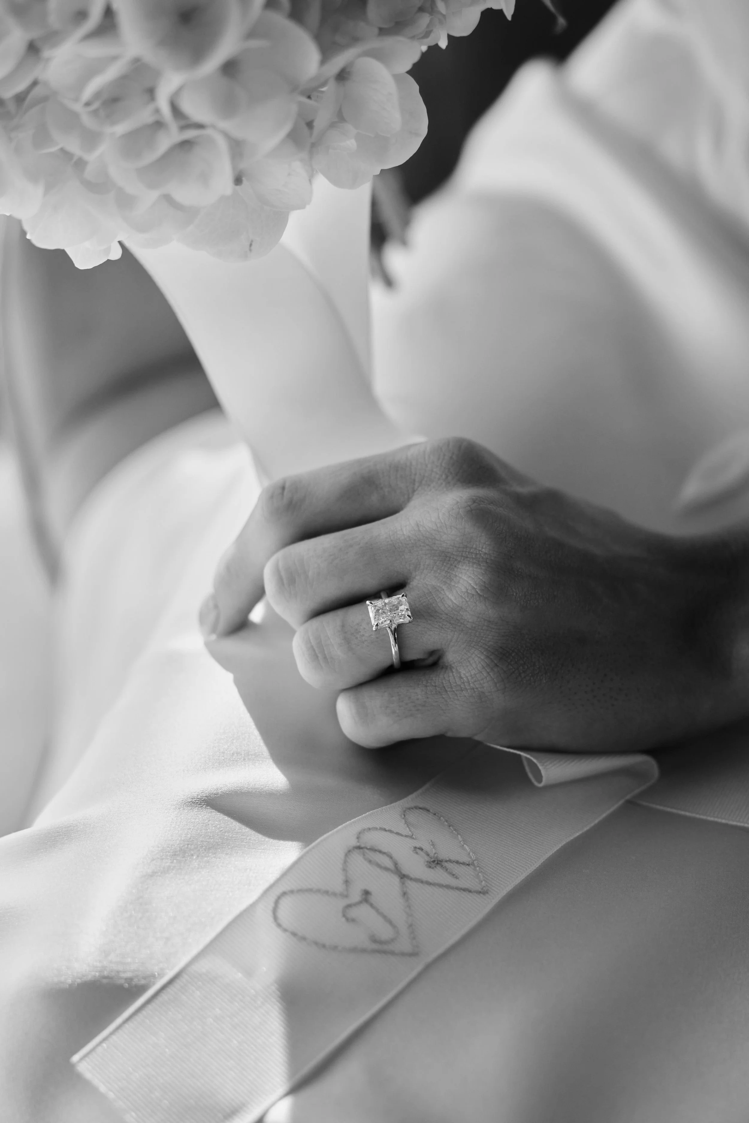 Close-up of a person's hand wearing a diamond engagement ring, resting on a satin fabric with a hand-stitched heart and initials, near a bouquet of flowers.