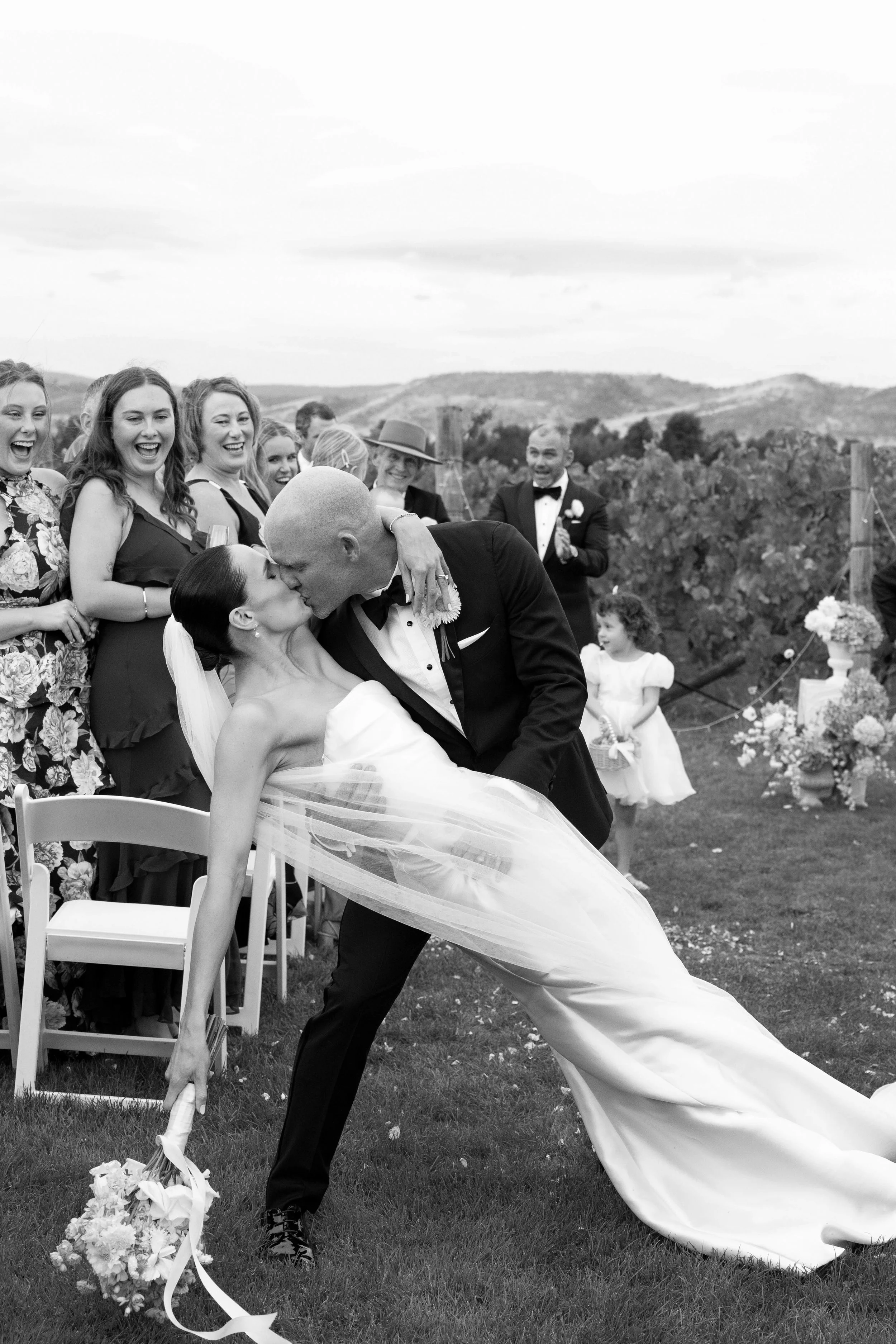 A couple on their wedding day share a kiss outdoors, with the groom dipping the bride. The bride is holding a bouquet, and guests are watching and smiling in the background, with a scenic landscape and rolling hills in the distance.