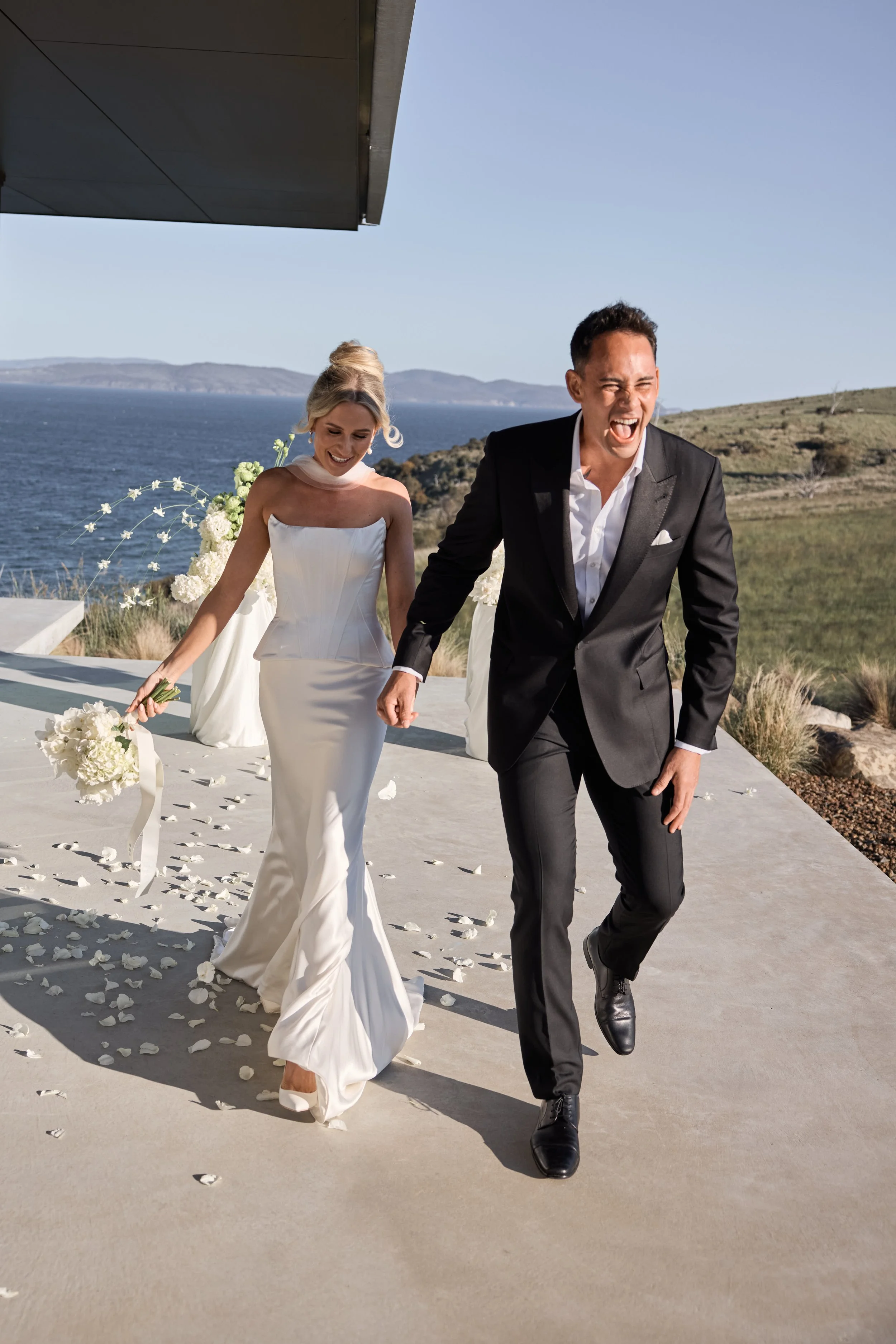 A bride and groom joyfully running hand in hand on their wedding day outside with a scenic ocean view in the background. The bride wears a white wedding gown holding a bouquet, and the groom is in a black tuxedo.