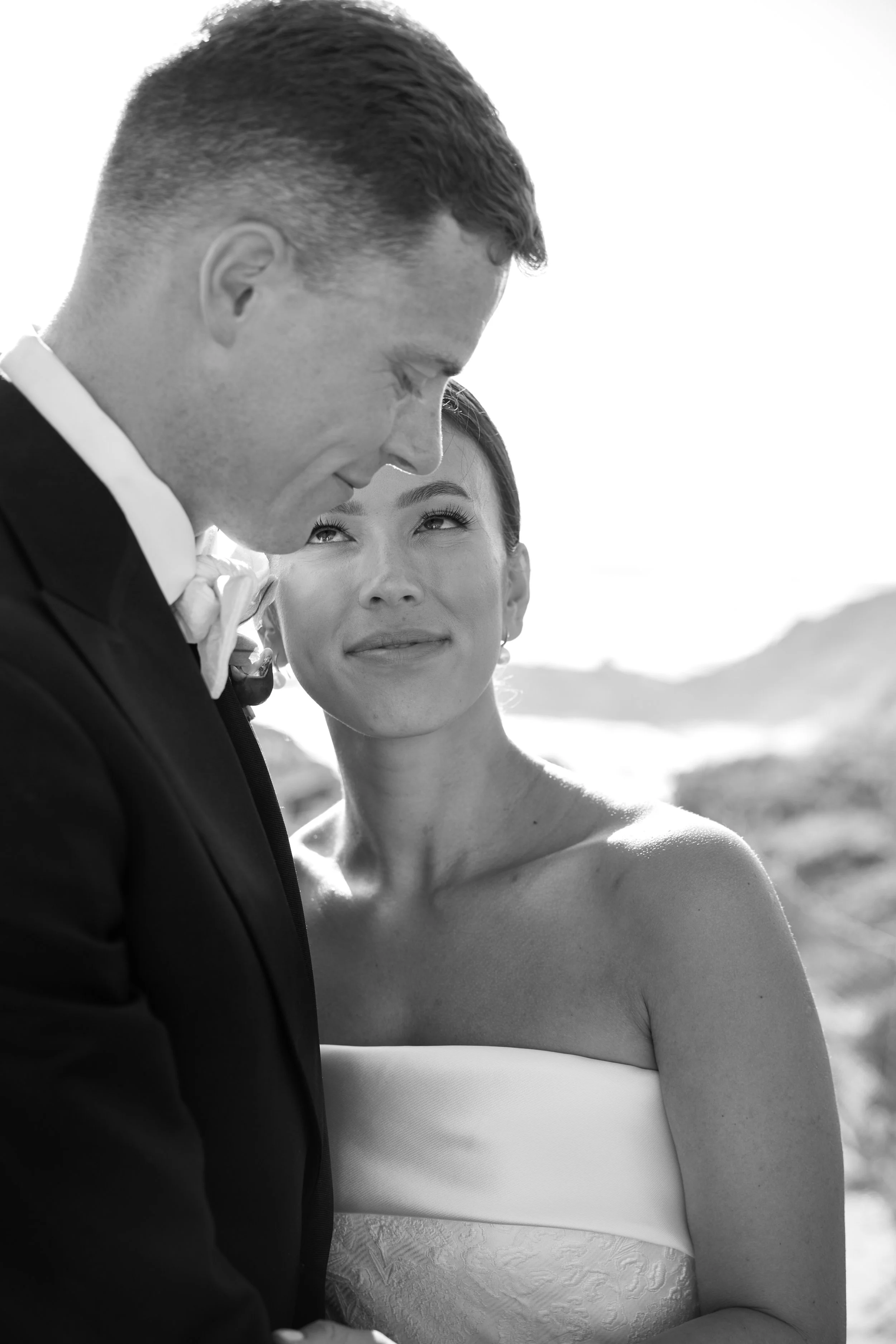 Black and white photo of a groom and bride outdoors, the groom leaning forward with eyes closed and the bride looking at him with a gentle smile, both dressed in wedding attire.