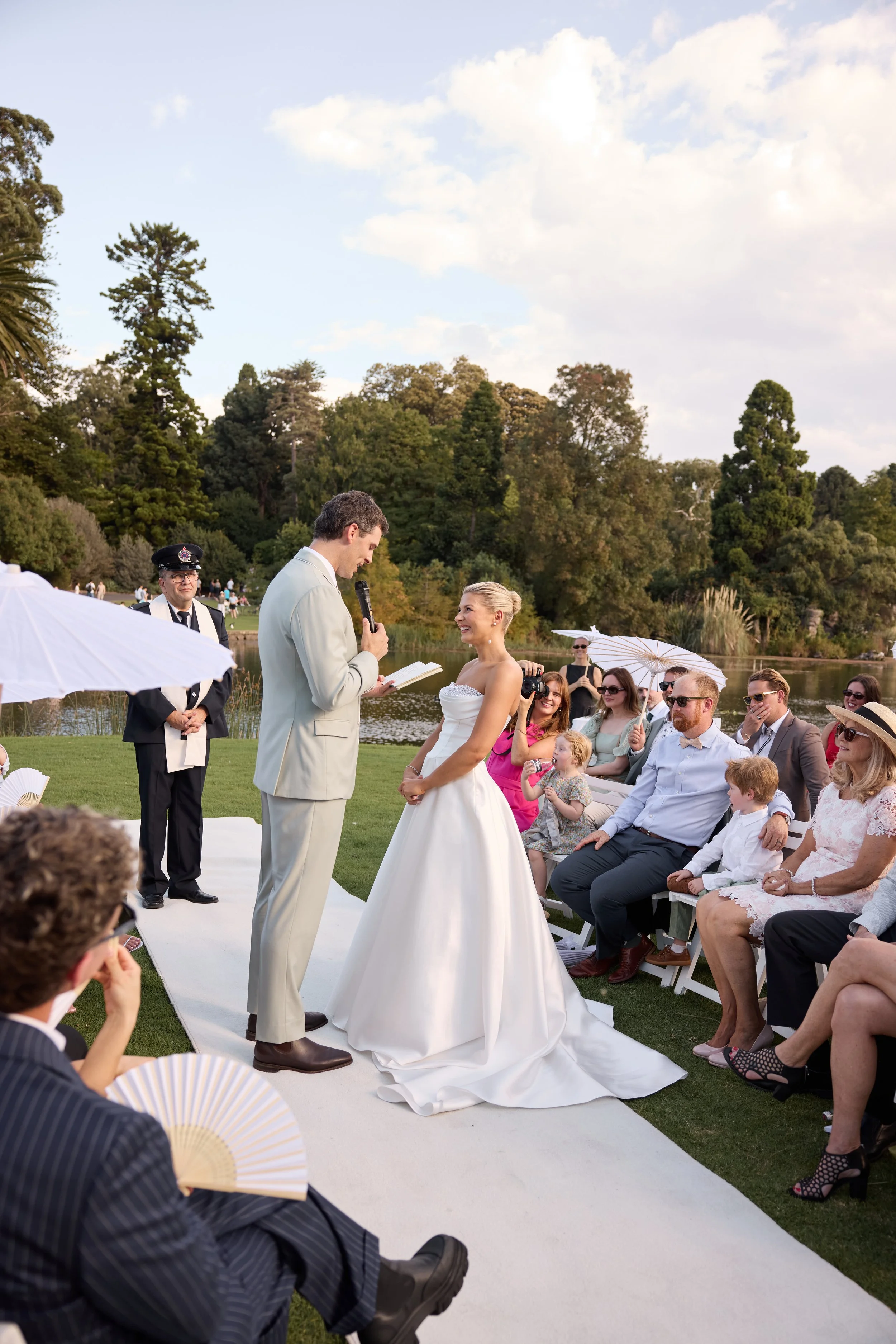 A wedding ceremony outdoors by a lake with guests sitting on chairs, the bride and groom standing under a sun umbrella, with the officiant reading from a book. The bride is smiling in a white wedding dress, and the groom is in a beige suit. People in