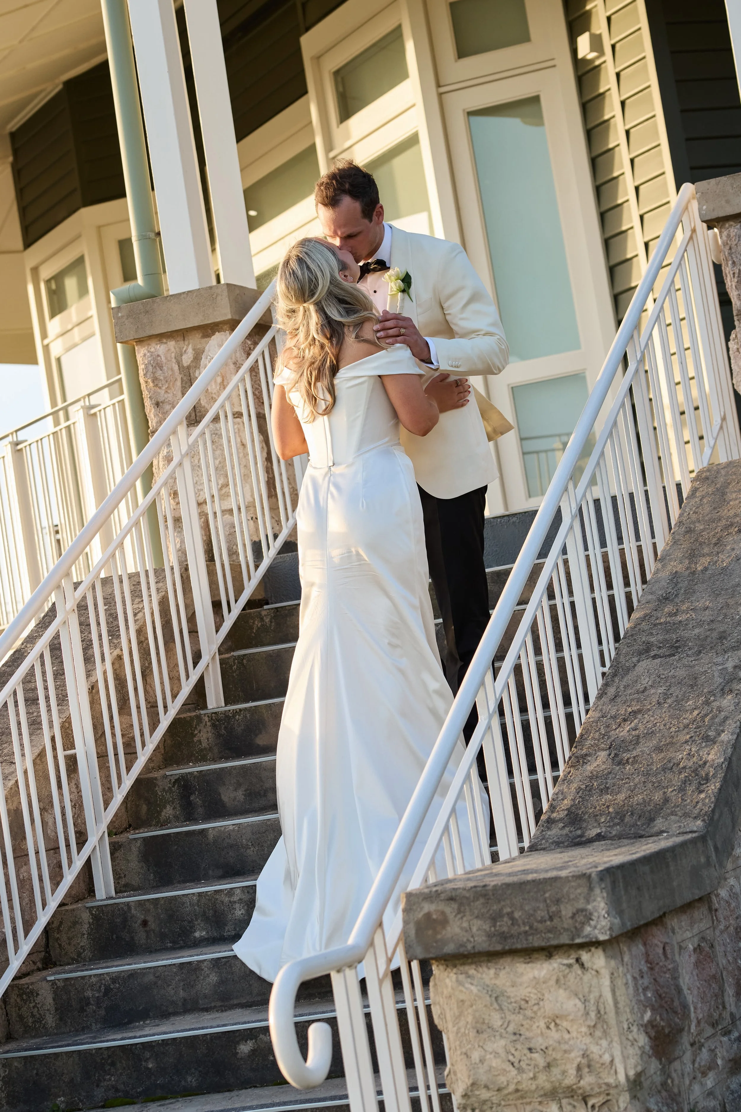 A bride and groom sharing a kiss on outdoor stairs, with the bride in a white wedding gown and the groom in a white tuxedo jacket, in front of a house with green siding.