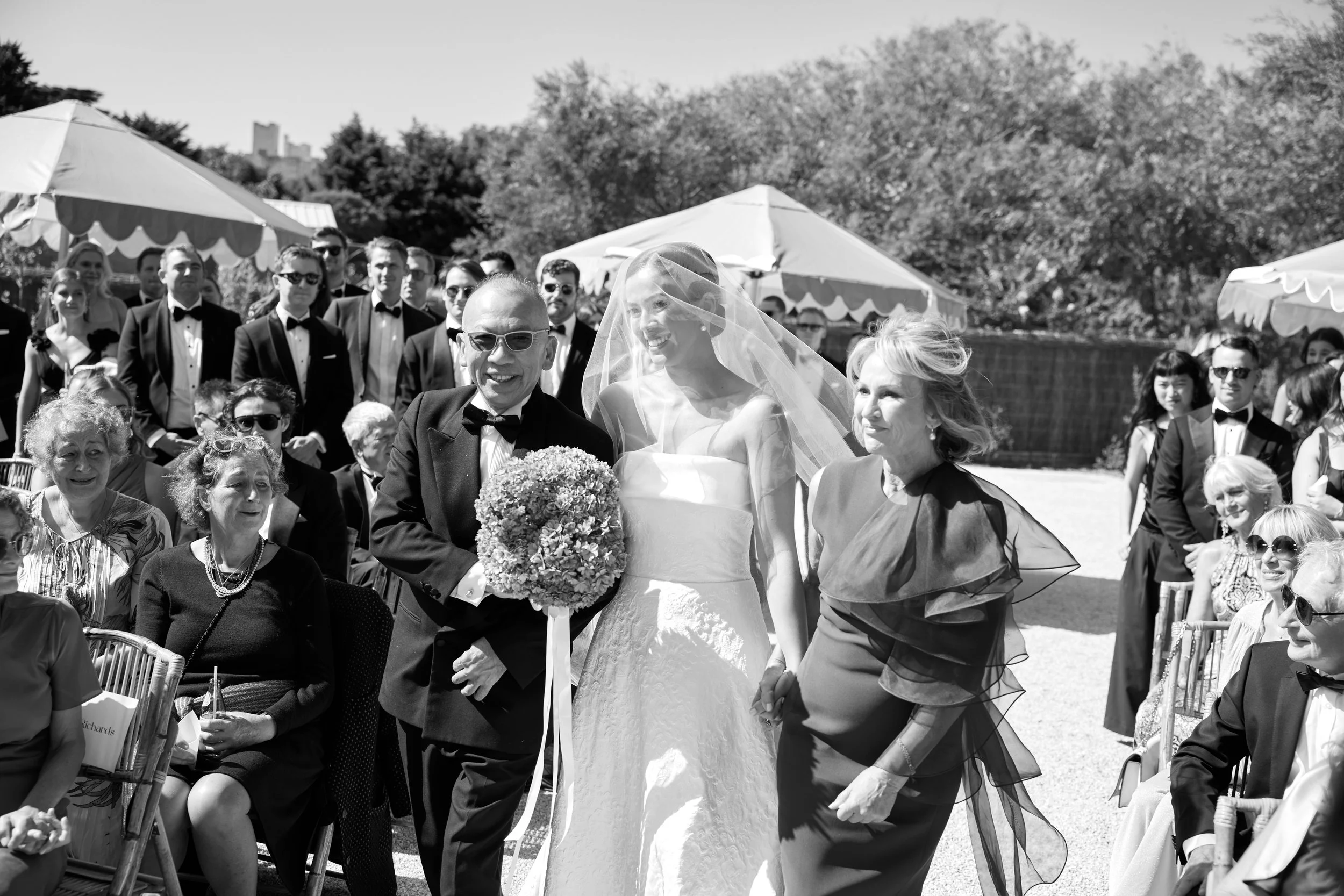 A black and white photograph of a wedding celebration outdoors with many guests dressed in formal attire, including tuxedos and dresses. The bride, wearing a white wedding gown and veil, is holding hands with an older woman. The groom, in a tuxedo, i