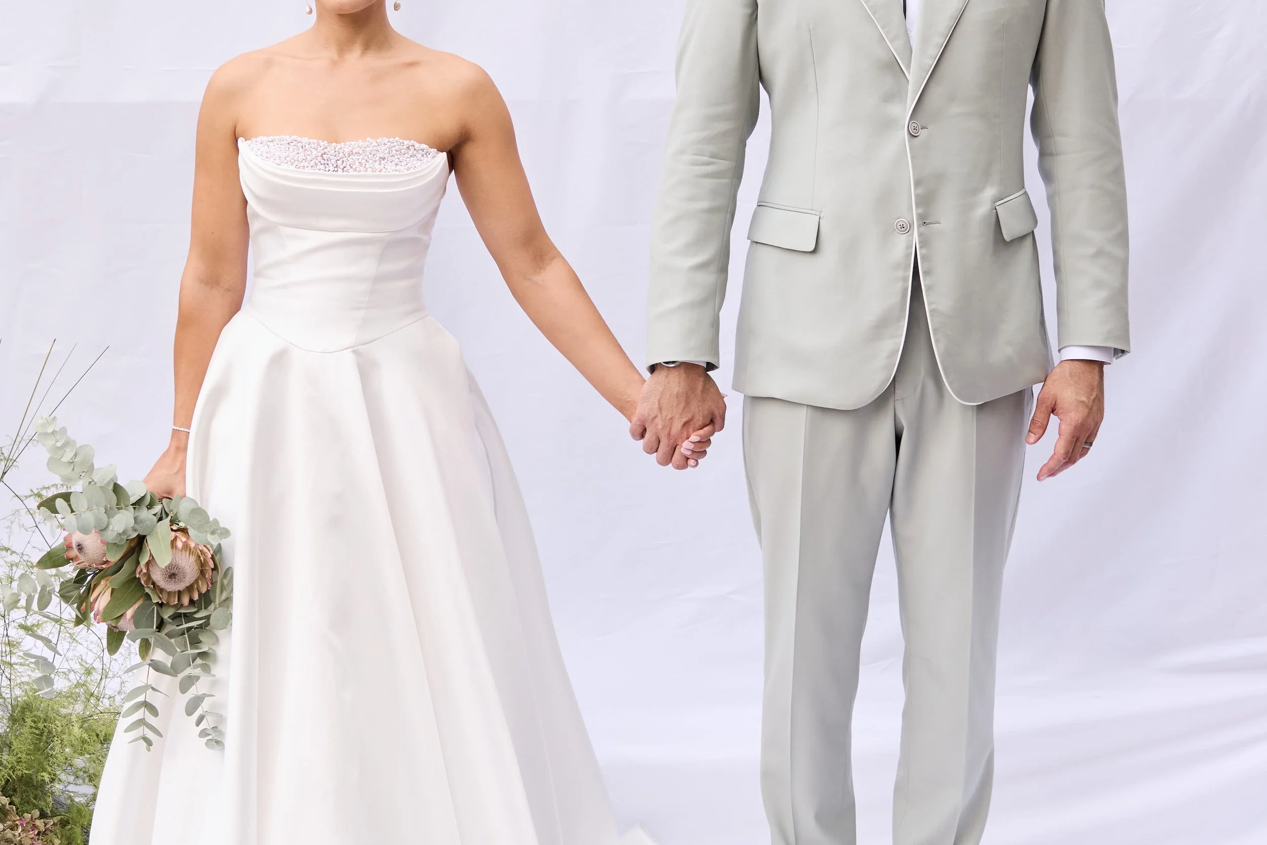 A bride and groom holding hands during their wedding ceremony. The bride is wearing a strapless white wedding gown and holding a bouquet of flowers. The groom is dressed in a light-colored suit.