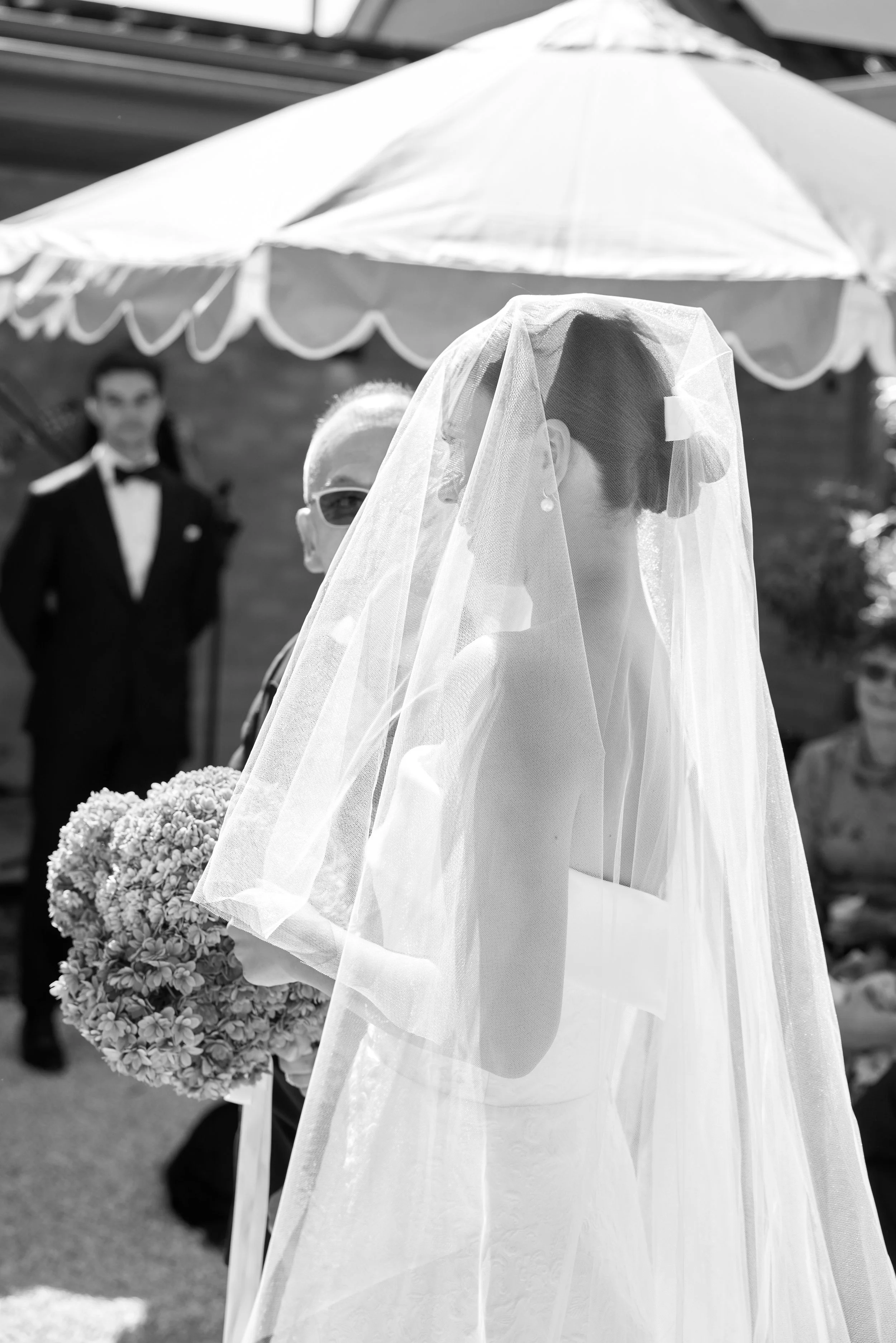 A bride with a veil holding a bouquet of flowers in an outdoor wedding ceremony. A man dressed as a groom is in the background.