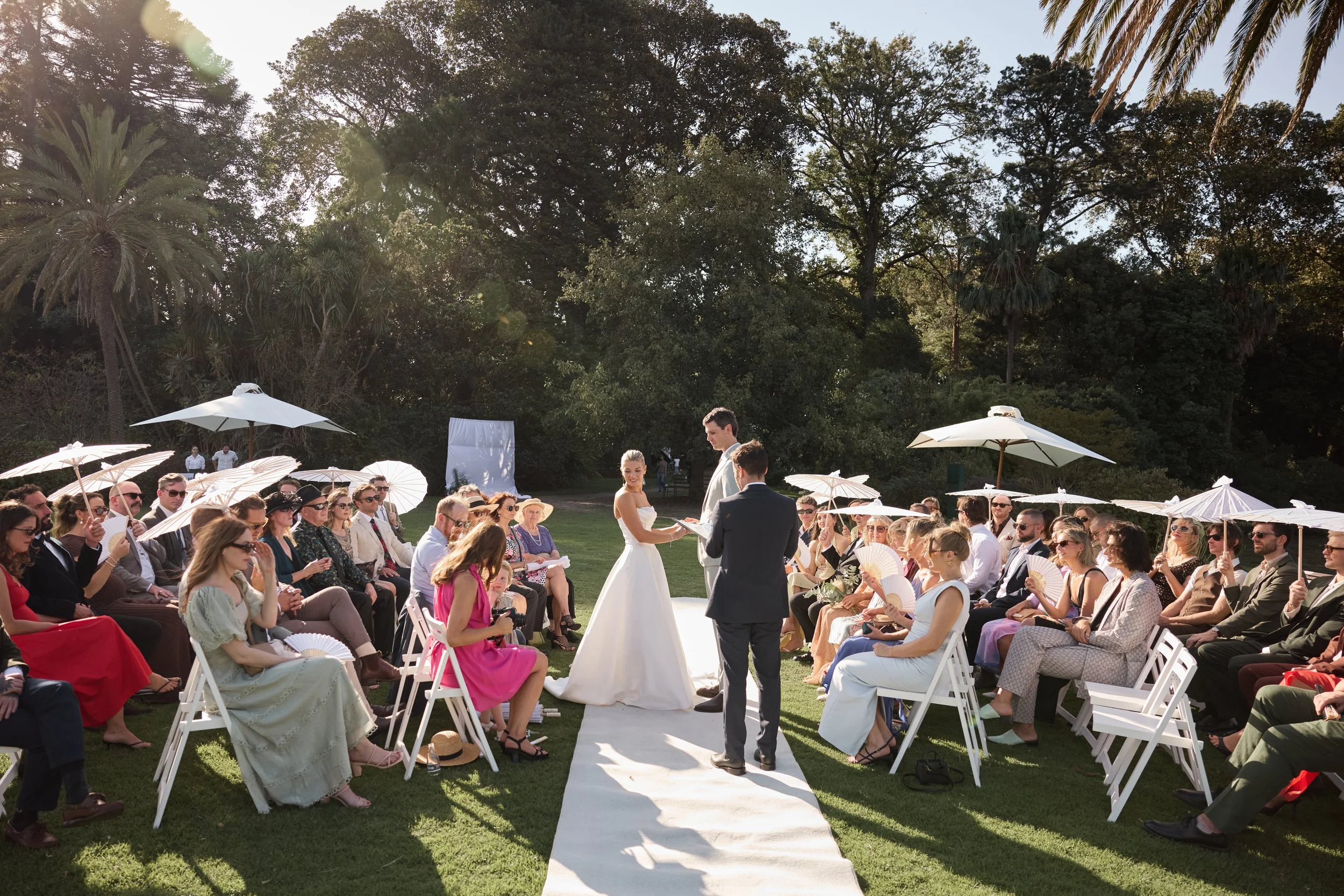 Outdoor wedding ceremony with guests seated under umbrellas, a bride and groom exchanging vows, set in a lush green garden with sunlight filtering through trees.