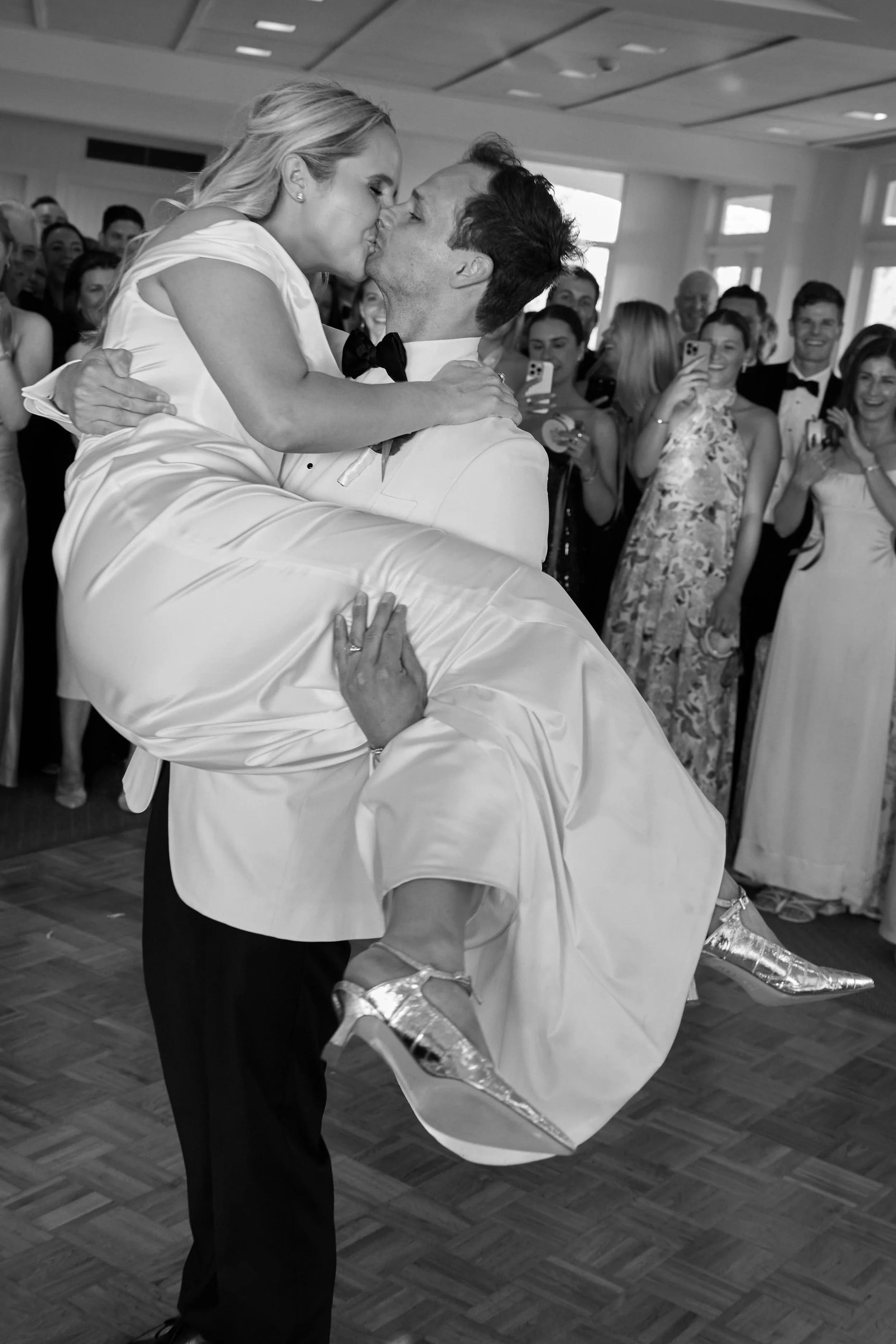 A man in formal attire lifting a woman in a wedding dress during a wedding reception, with guests watching and taking photos in the background.